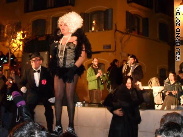cross dresser: spain Alicante Carnival Fiesta, Costa Blanca, darkened plaza, bright-lit stage, spectators dressed in black and red, black man dance, skimpily dressed singer in drag, musicians