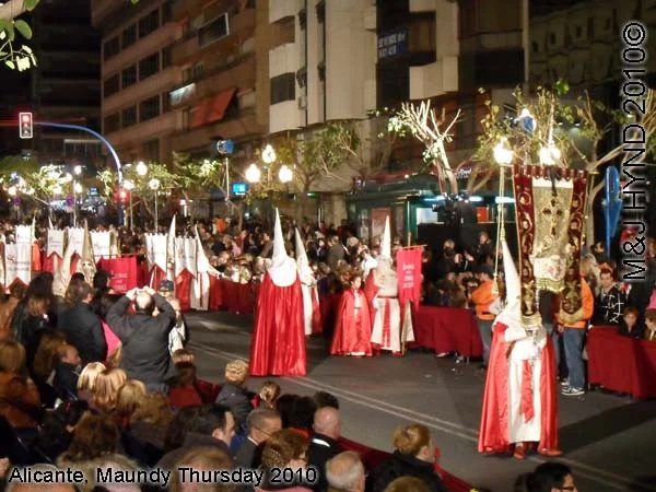 pain Alicante, Semana Santa Holy Week, Maundy Thursday city procession, Brotherhood long pointed white hoods, long capes carry lamps on poles, flag-bearers, marching bands