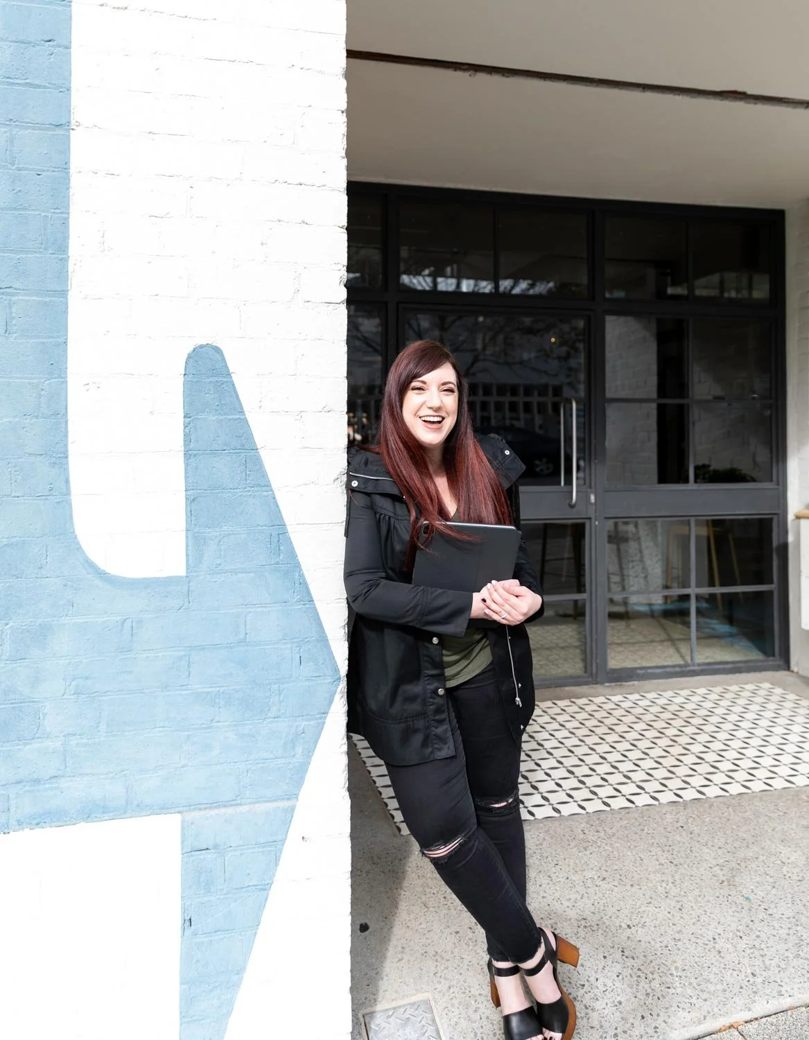 Joanne Tapodi, Australian Brand Strategist and Squarespace Website Designer leaning against a wall holding iPad and laughing