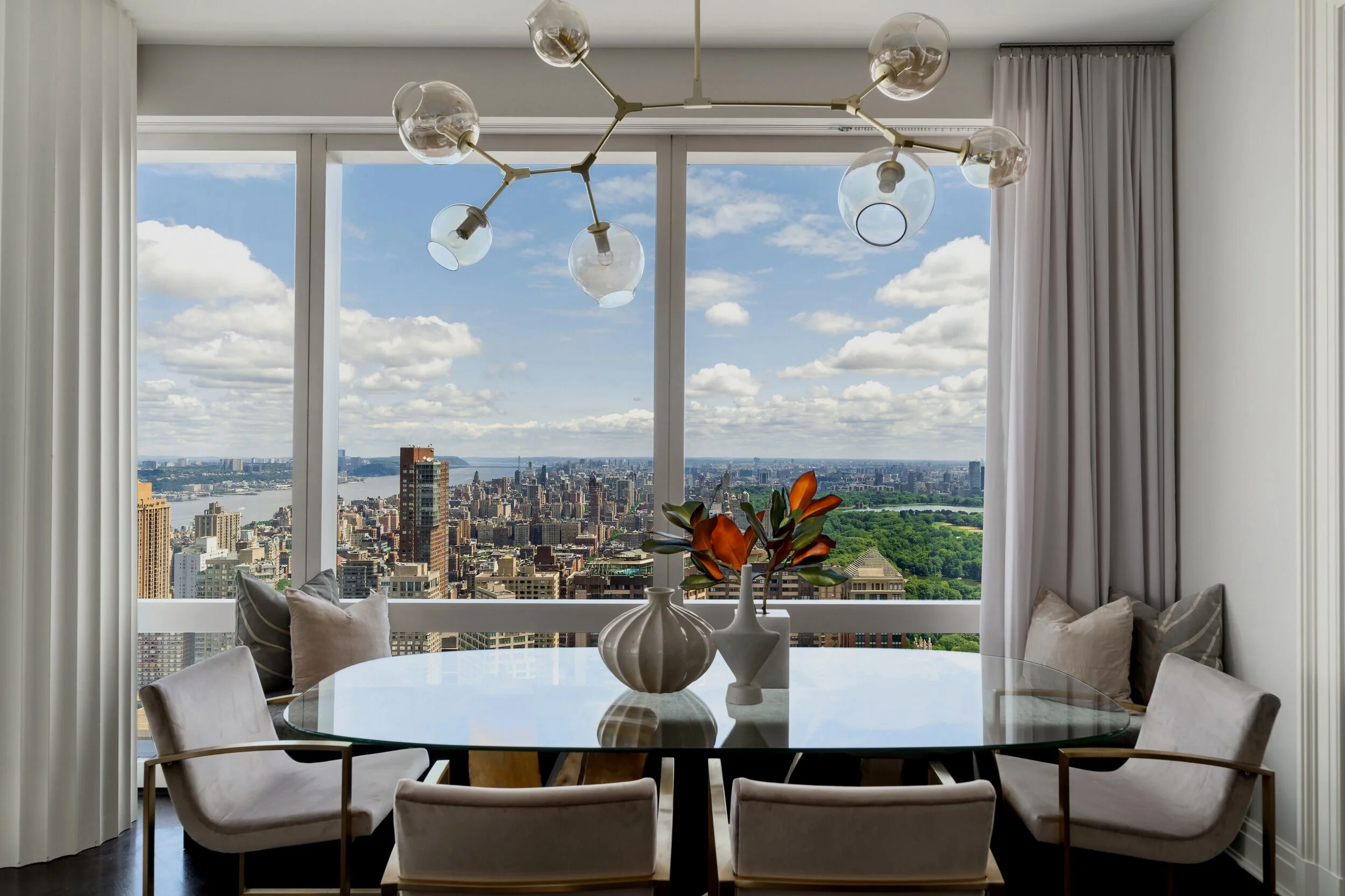 Modern dining room with a circular glass table surrounded by white chairs and cushions, a white vase with orange and green flowers, large windows with curtains revealing a city skyline and green park, contemporary chandelier with multiple glass globes.