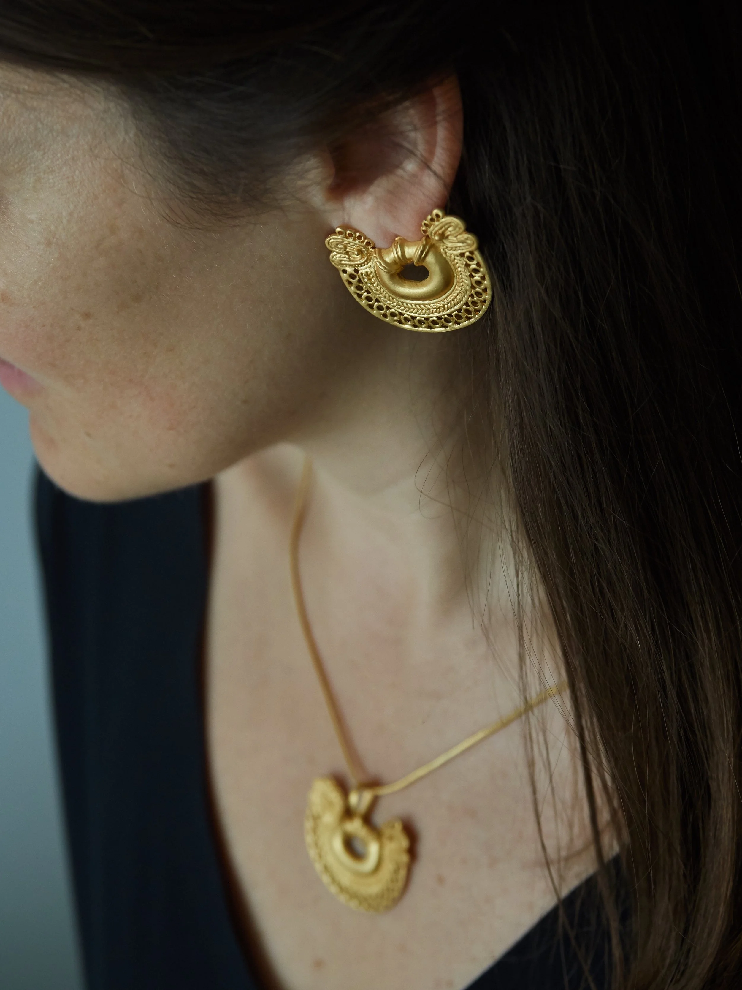 Gold-plated Tairona-inspired double-headed serpent earrings worn on the ear, based on pre-Columbian Colombian goldwork.