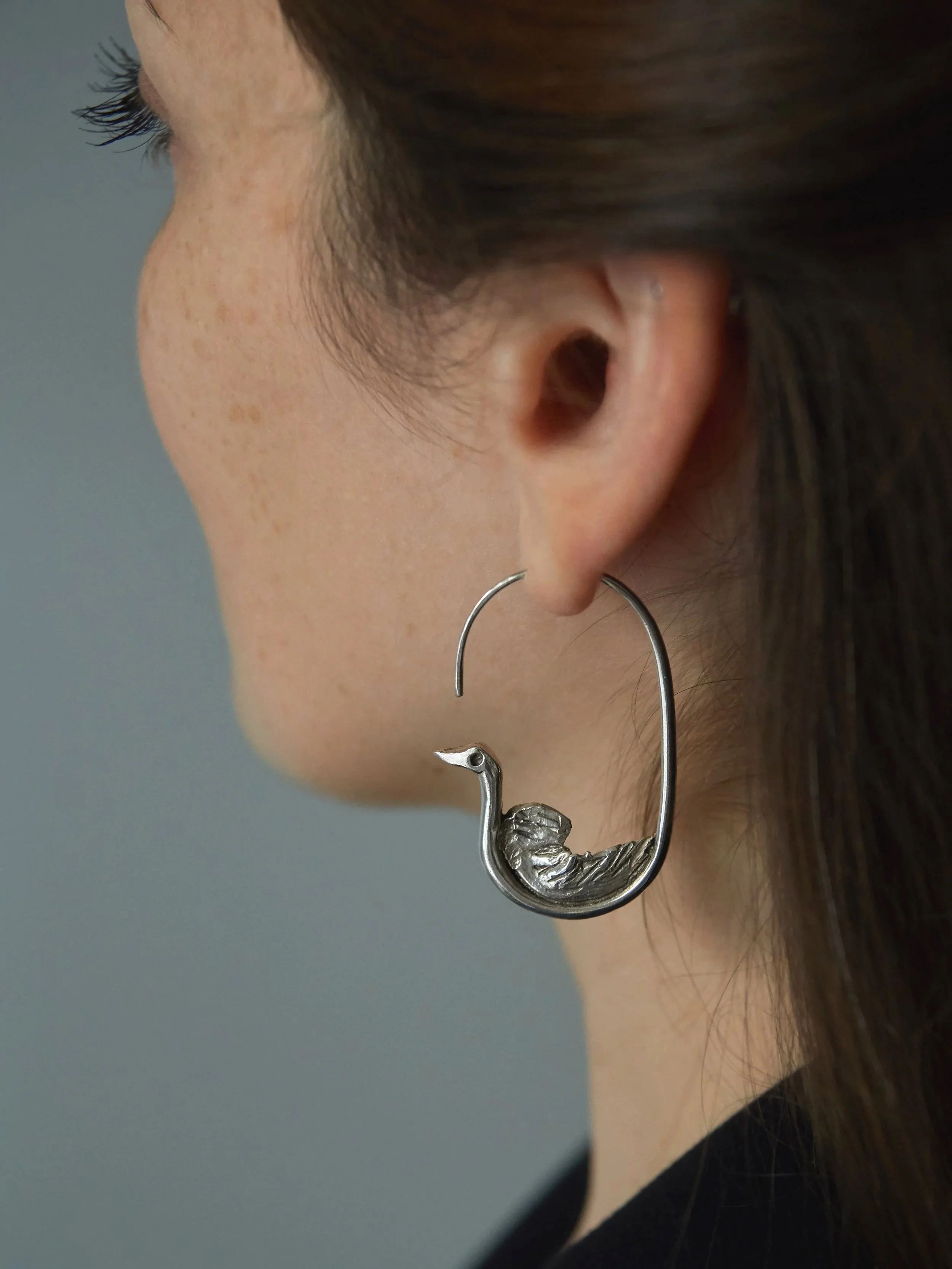 Elisabeth Riveiro’s Oiseau de l’Arrivée earrings in silver-plated bronze, featuring a sculptural bird motif with textured wings and an elongated open-hook design, worn on the ear.