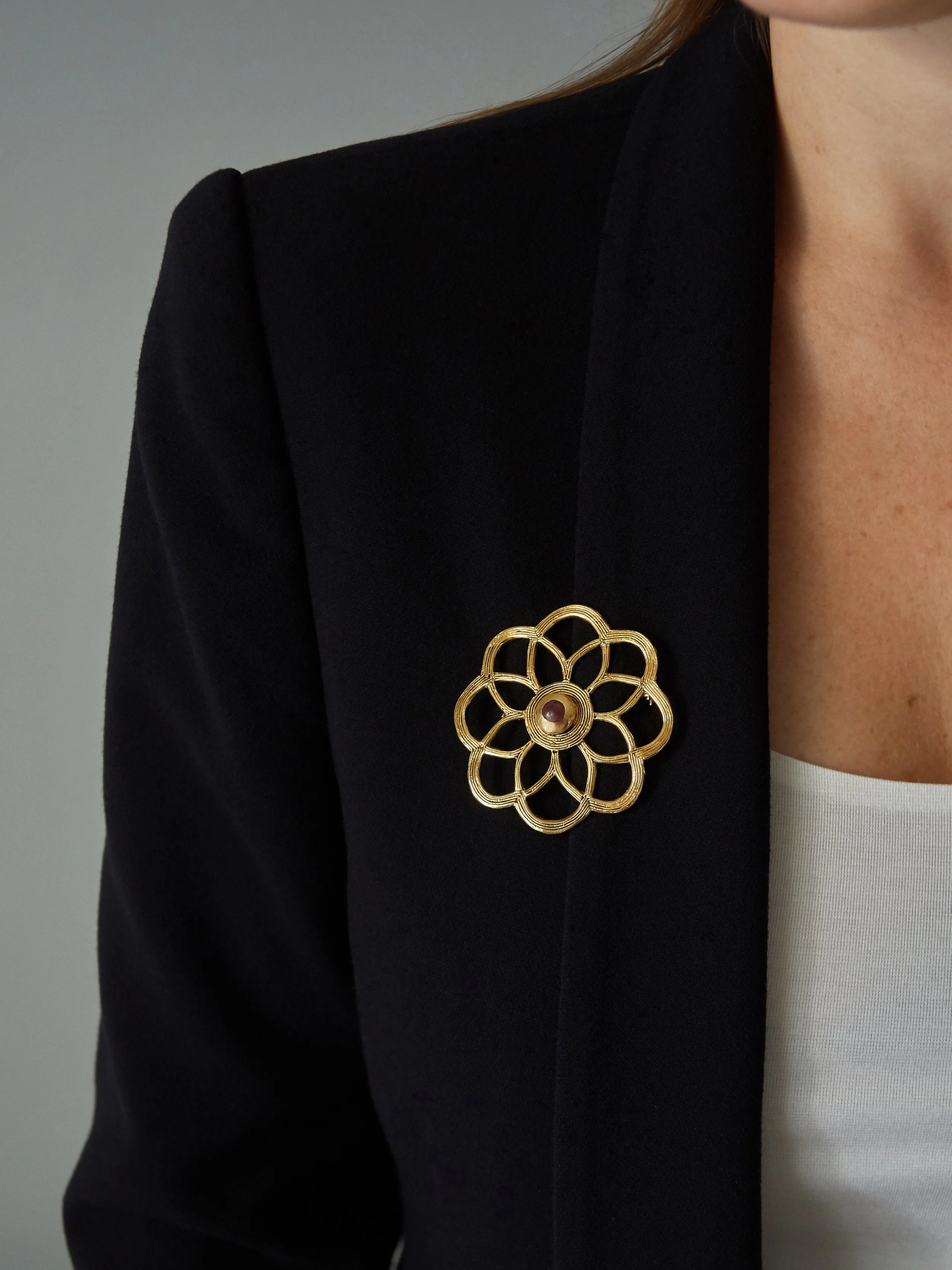 Elisabeth Riveiro’s Jaïpur floral brooch in gold-plated bronze, featuring an openwork geometric flower design with a red enamelled centre, worn on a black blazer.