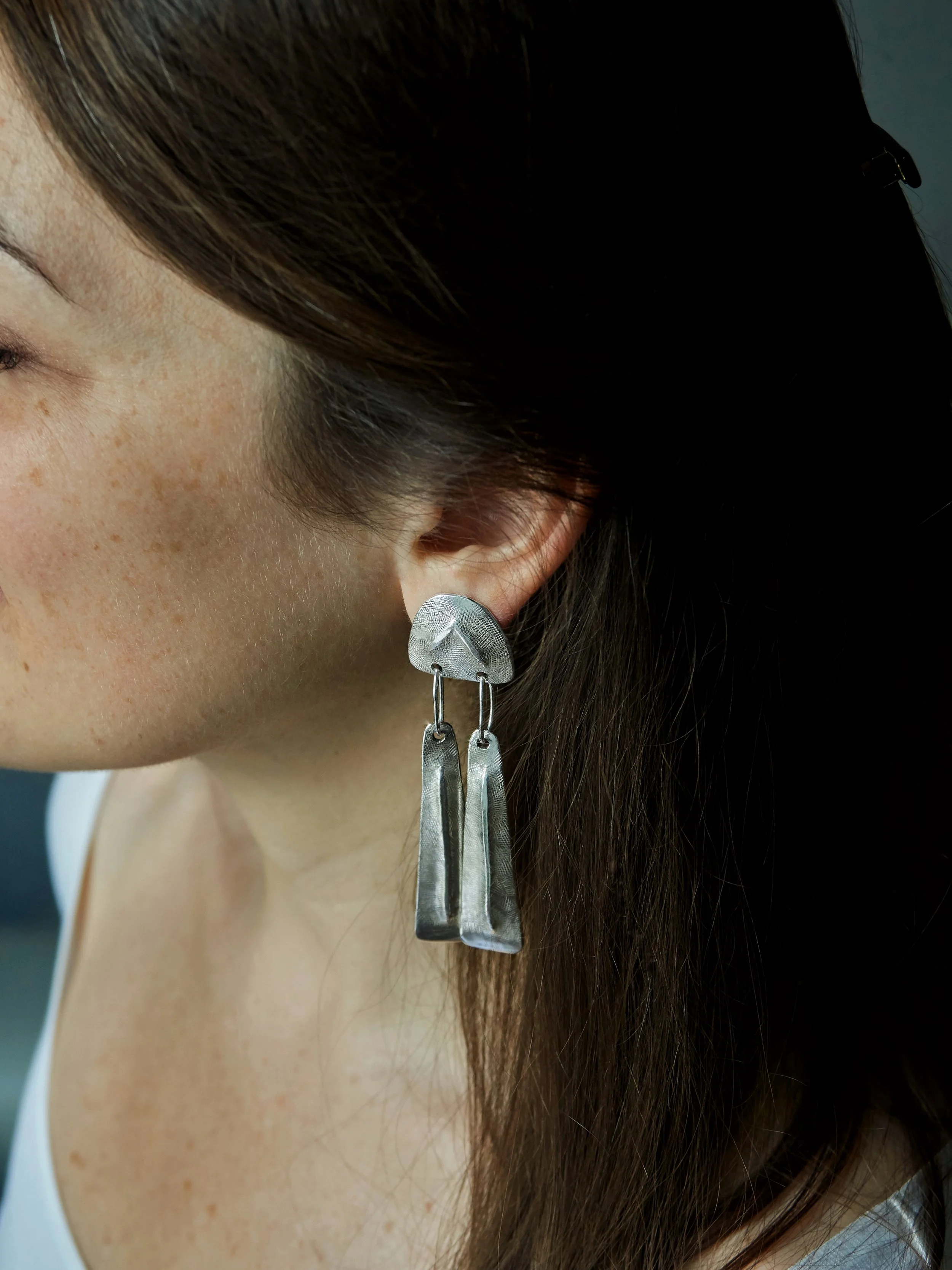 Elisabeth Riveiro Vénus long earrings in silver-plated bronze, worn to reveal their architectural silhouette, textured metal surfaces and two freely suspended pendants. Handmade in Paris.