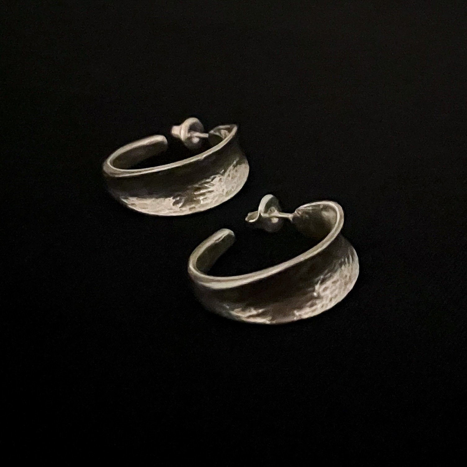 Pair of Petit Val silver-plated bronze hoop earrings by Elisabeth Riveiro, photographed against a black background. Sculptural design with a hammered surface.