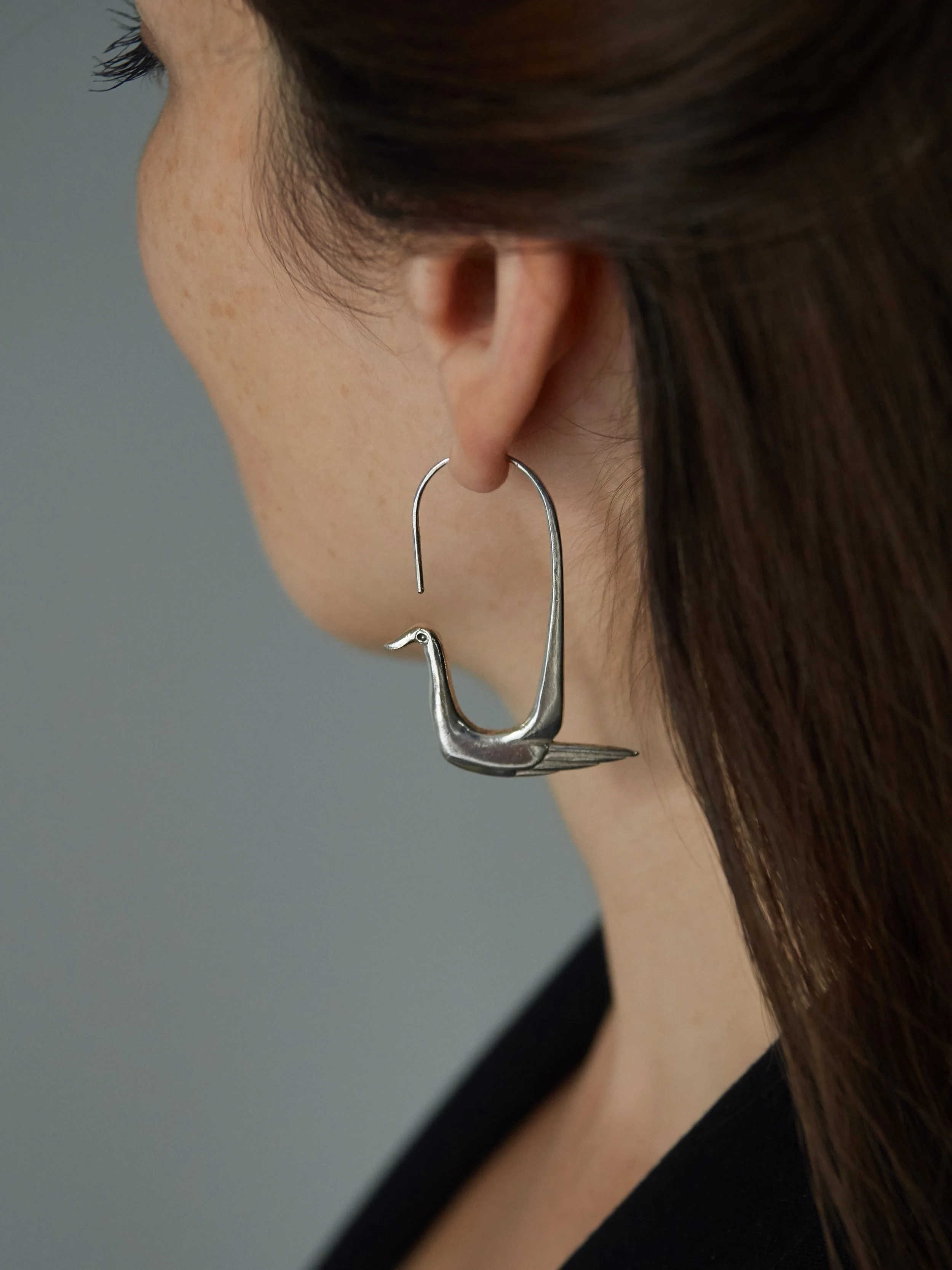 Elisabeth Riveiro’s Super Canard earrings in silver-plated bronze, featuring a stylised duck with an elongated neck, geometric body and sculptural open-hook design, worn on the ear.