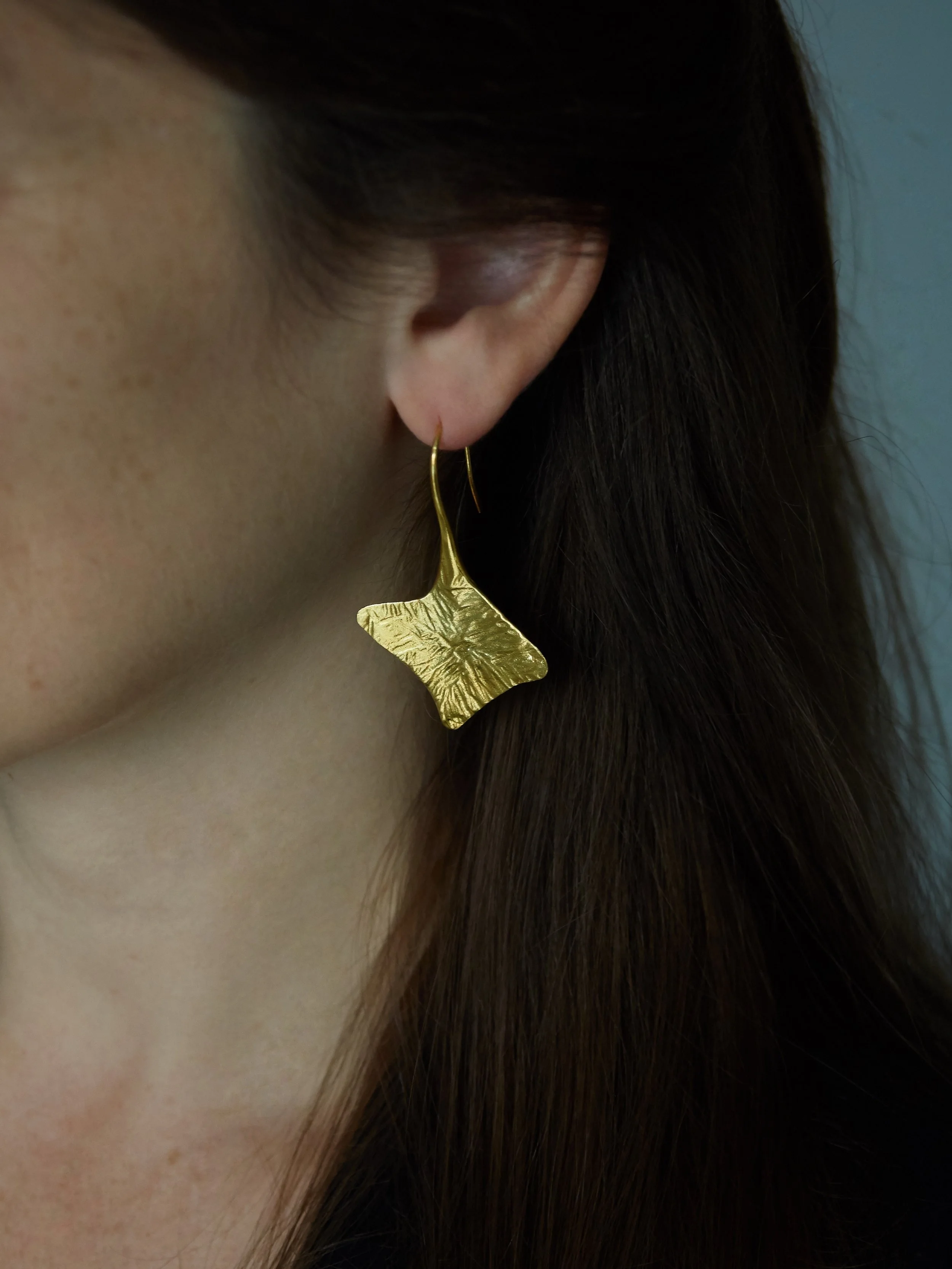 Palmyre gold-plated bronze earrings by Elisabeth Riveiro, featuring an asymmetrical organic shape with radiating relief patterns and an elongated ear hook.