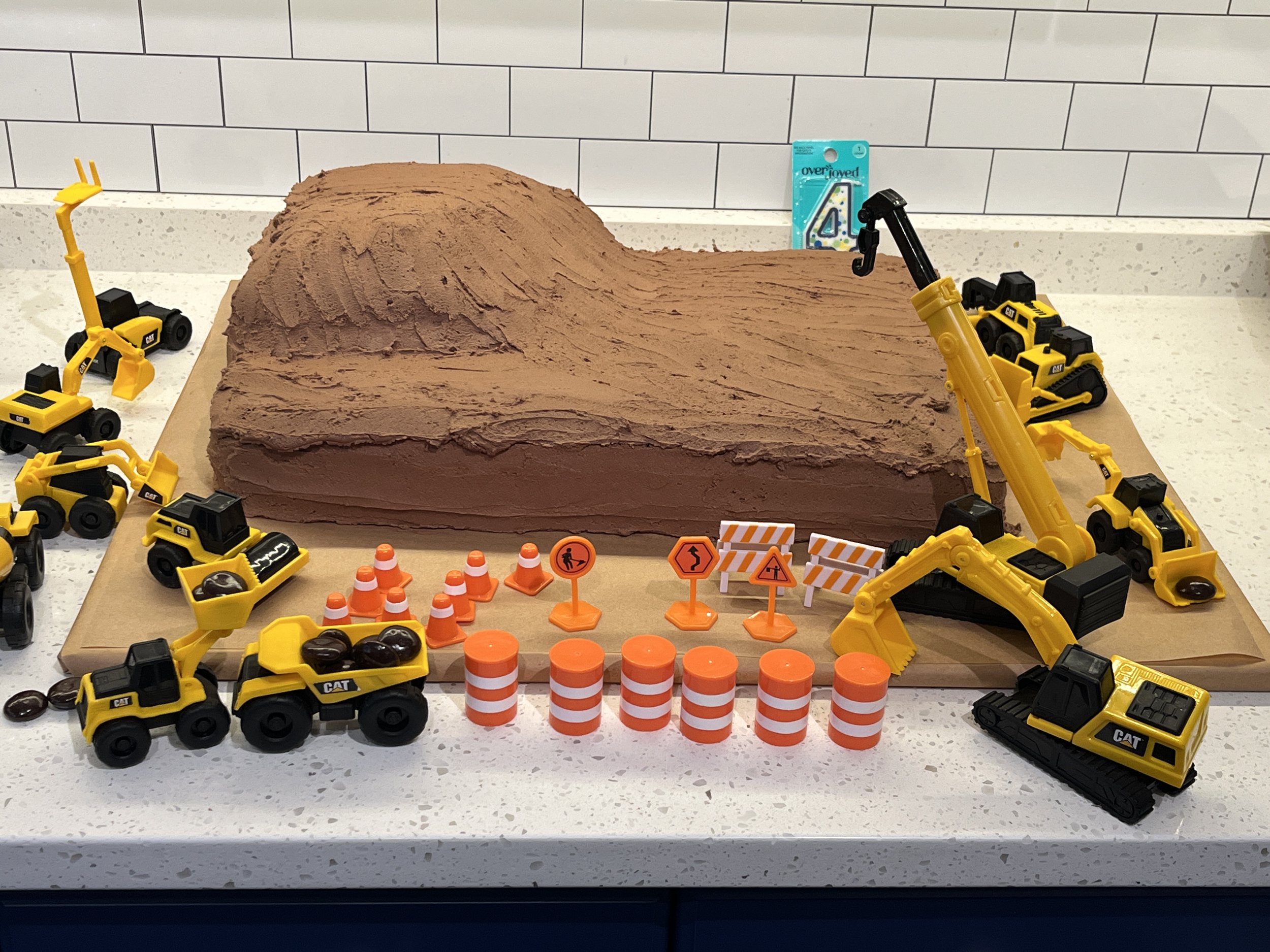Frosted construction zone cake with the final top decor set around it. Plastic cones, and construction equipment designed to be cake toppers are staged before final placement.
