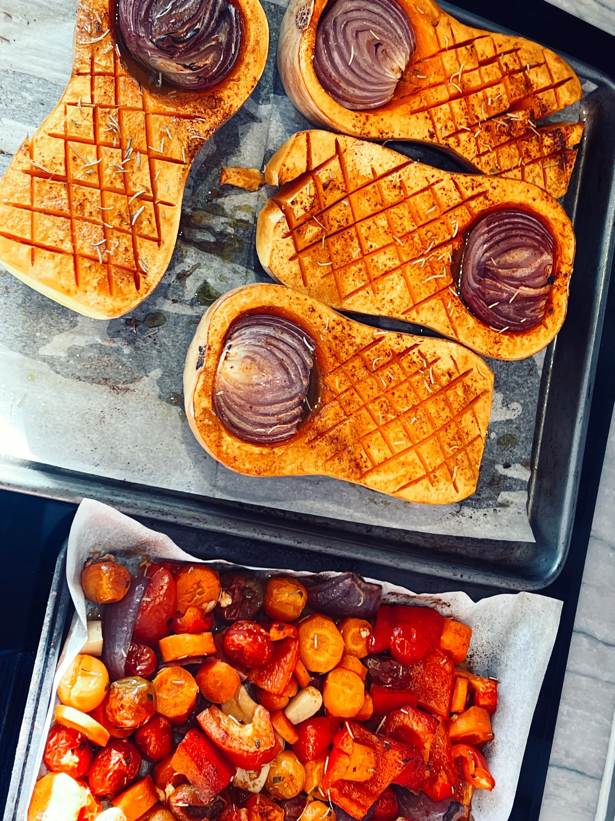 Roasted Butternut Squash Vegetable Soup being prepared on an oven roasting sheet. Onions are shown roasting inside butternut squashes. Also on the roastin sheet pan are colorful bell peppers, carrots, tomatoes, and red onion.