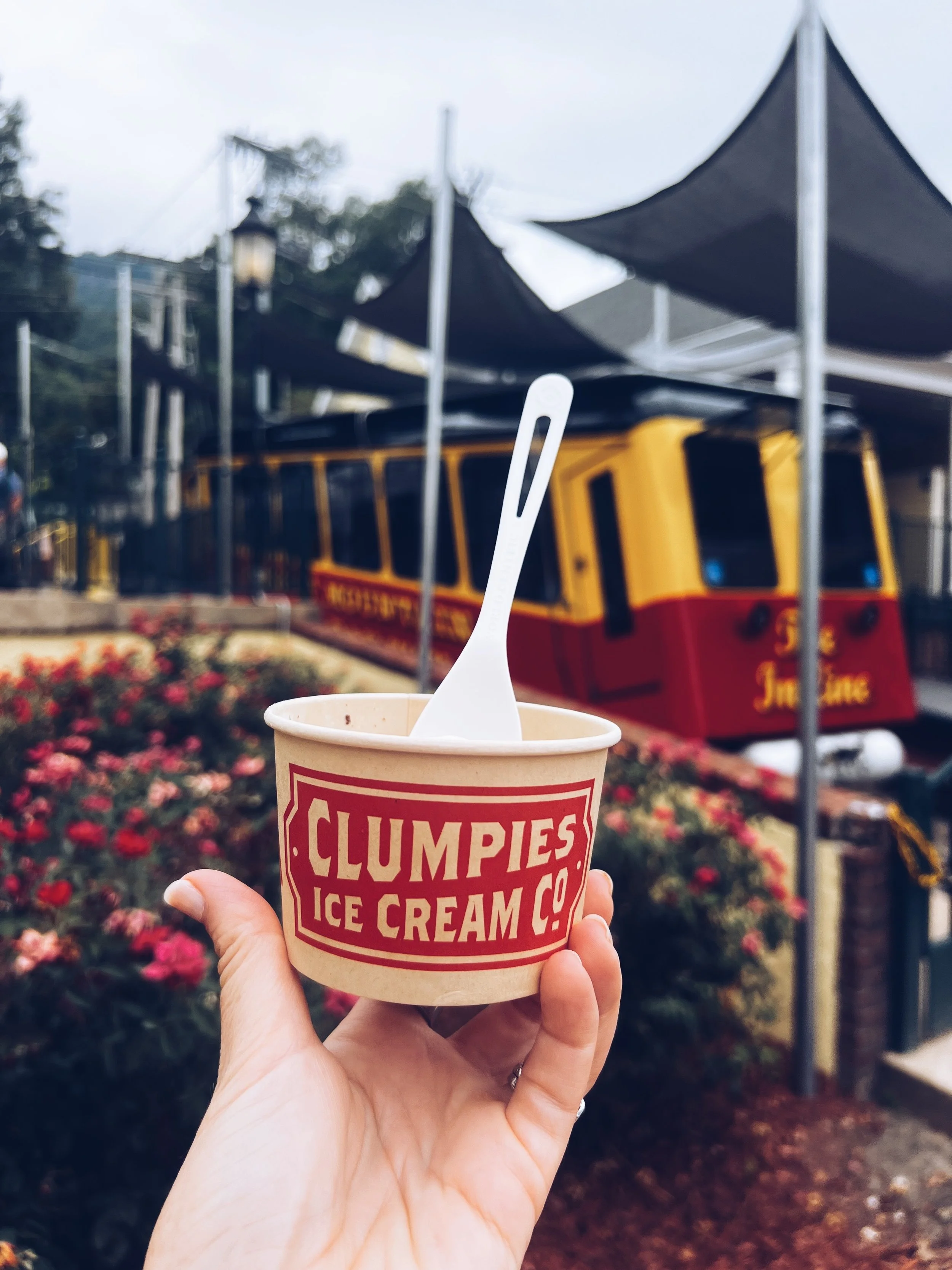 Clumpies Ice Cream Chattanooga, TN. Picture has a hand holding a cup of in-house made ice cream with the Incline Railway in the background.