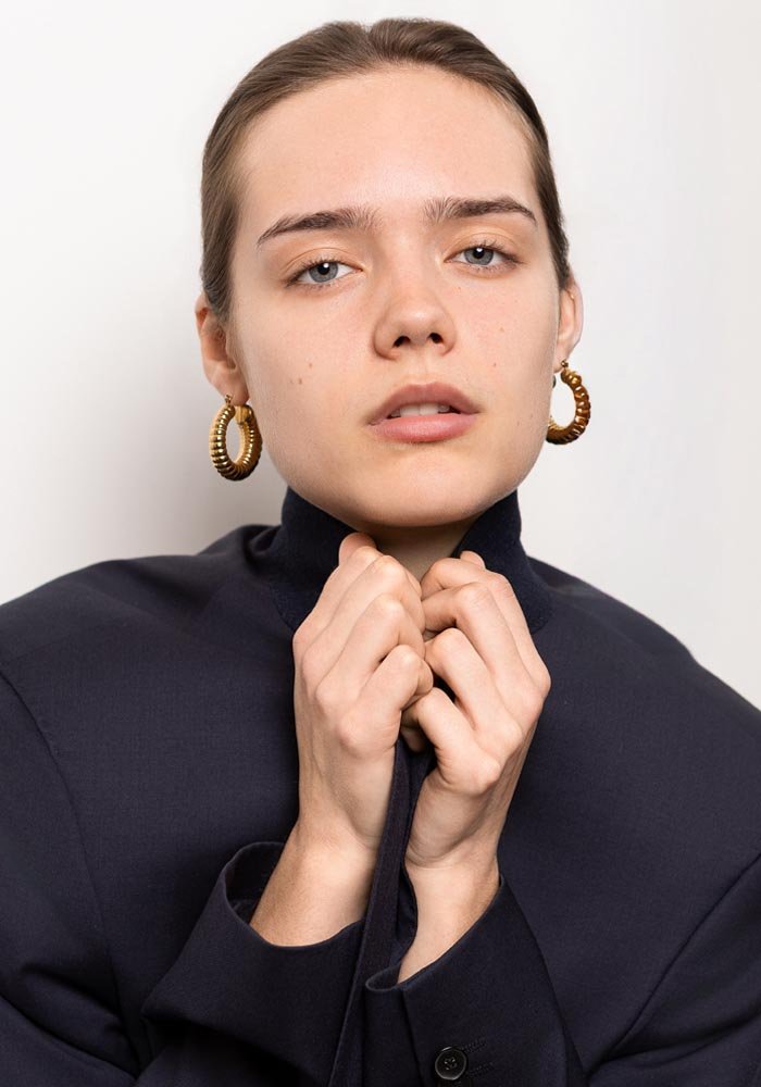 Jeune femme avec des boucles d'oreilles or, portant un haut noir, posant devant un mur blanc.