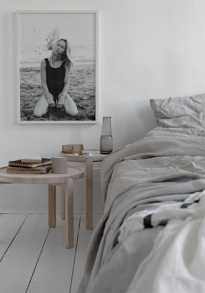 Une chambre minimaliste avec un lit non fait, une table de chevet en bois clair avec des livres, une bougie et une carafe en verre, et un tableau noir et blanc d'une femme assise sur la plage.