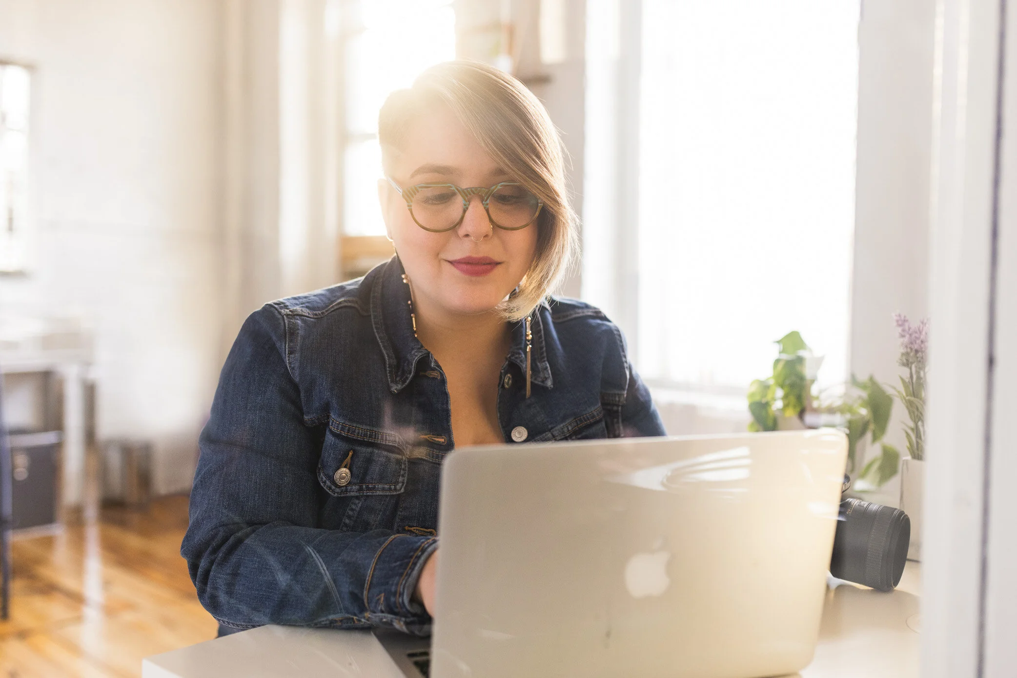 Quirky, cute gal sits in front of their computer in a jean jacket with light spilling in the window behind them, emailing clients.