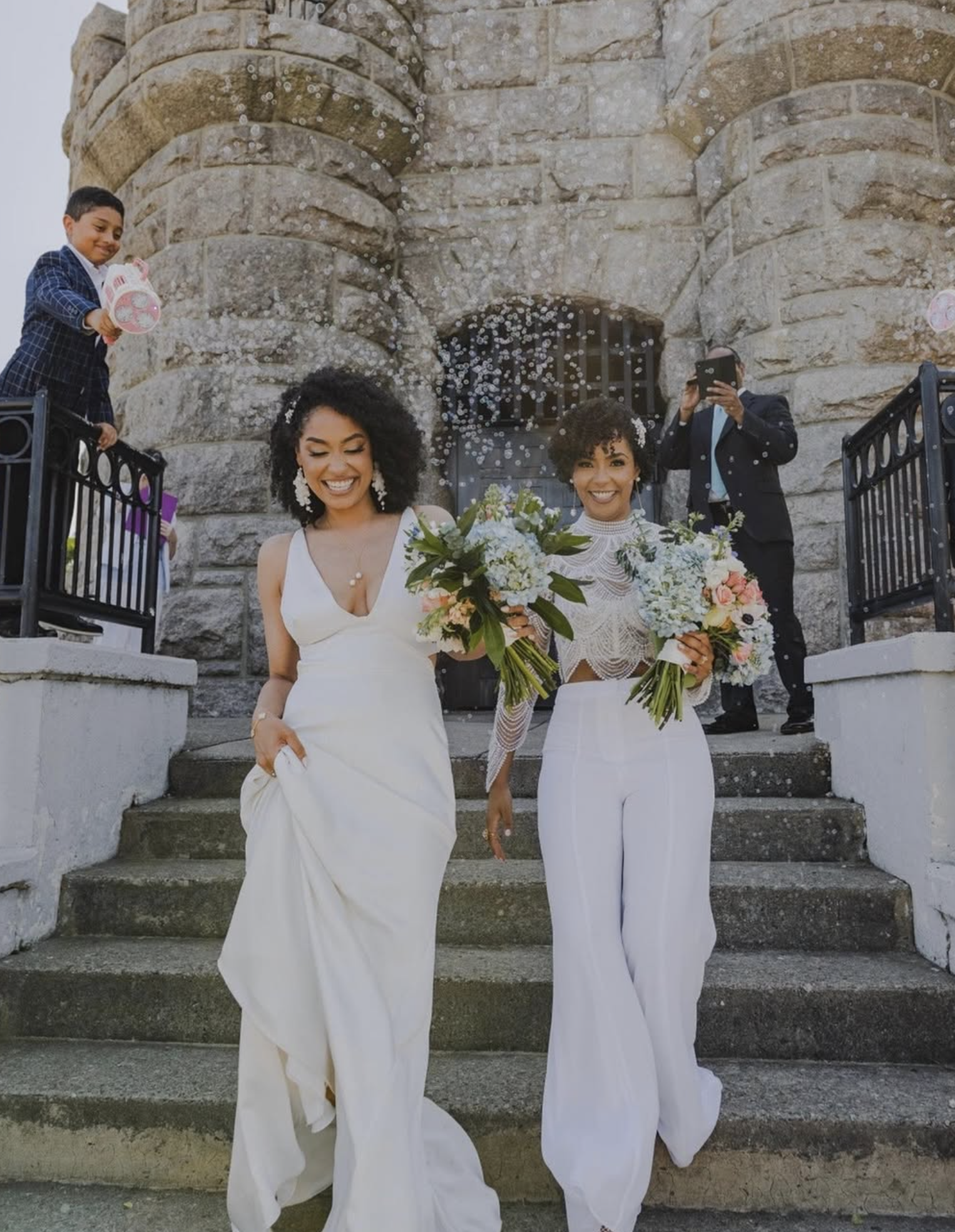 Two brides in white holding bouquets grin joyfully as they process down the stairs of Prospect Hill Park's tower, surrounded by family, friends, and bubbles after just being pronounced married.