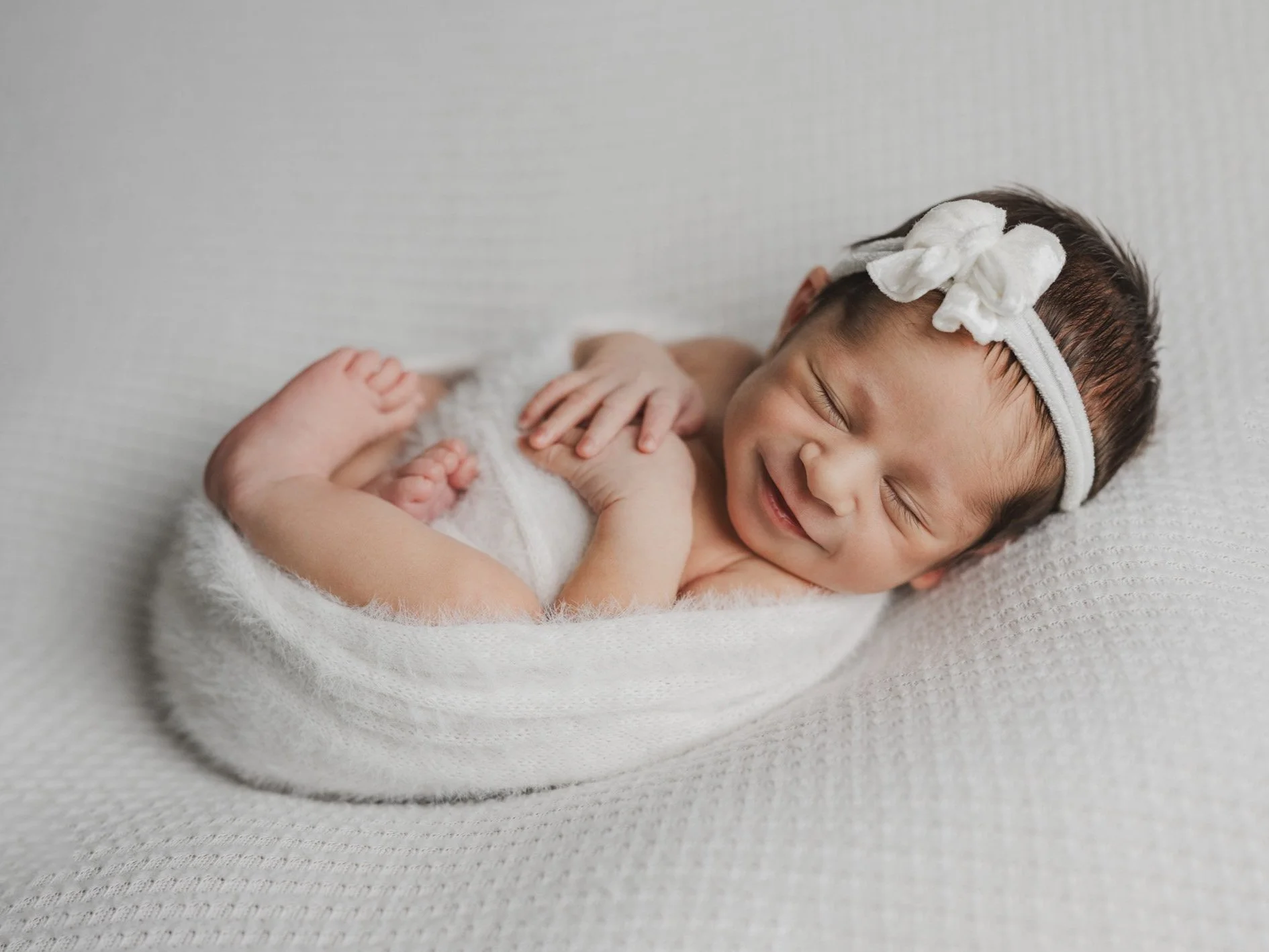 Smiling newborn girl in a white velvet bow, swaddled in white fabric with her feet and arms crossed lays on her back on white fabric.