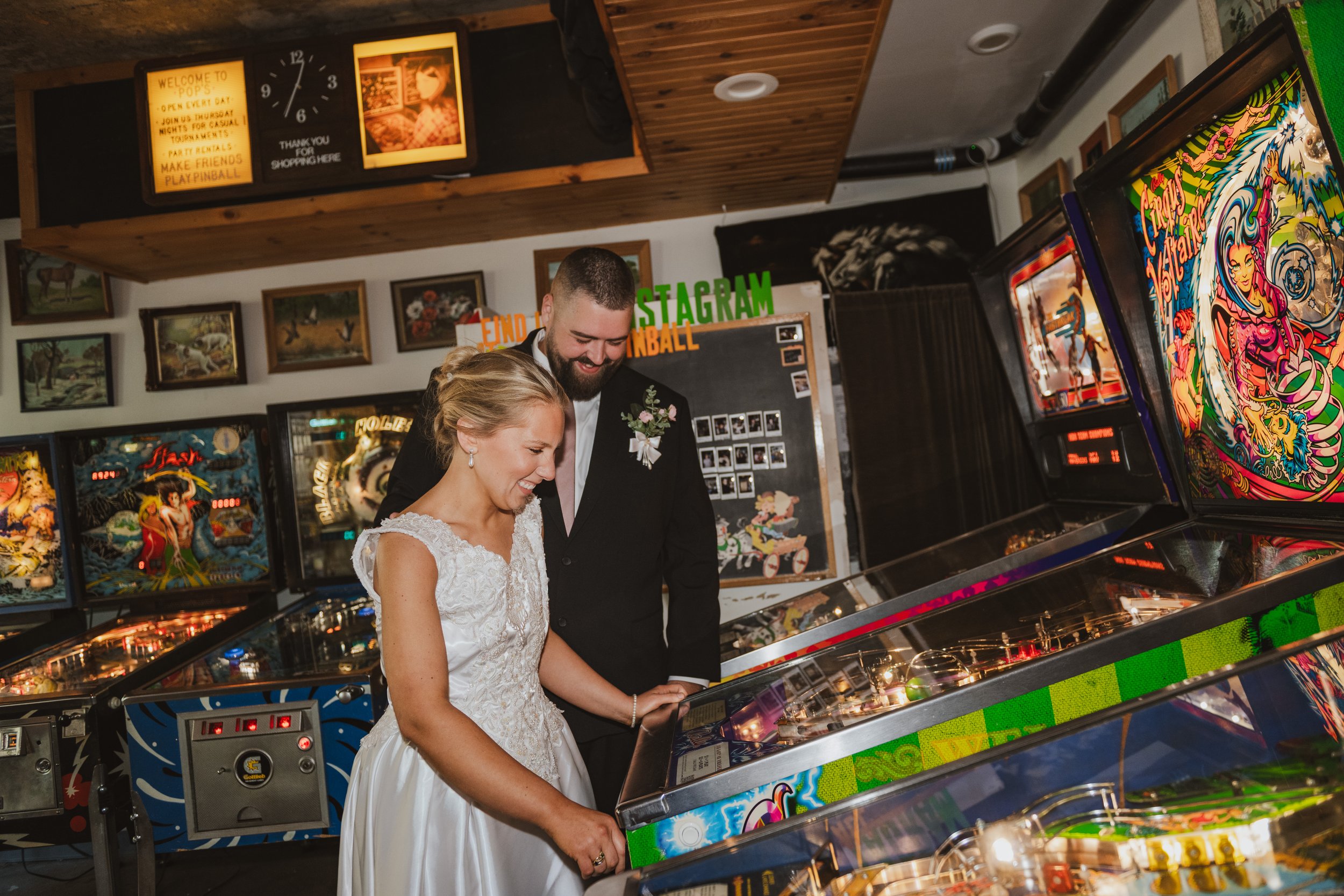 A couple in wedding attire laughs while they play pinball after being pronounced husband and wife at Dearly Studio