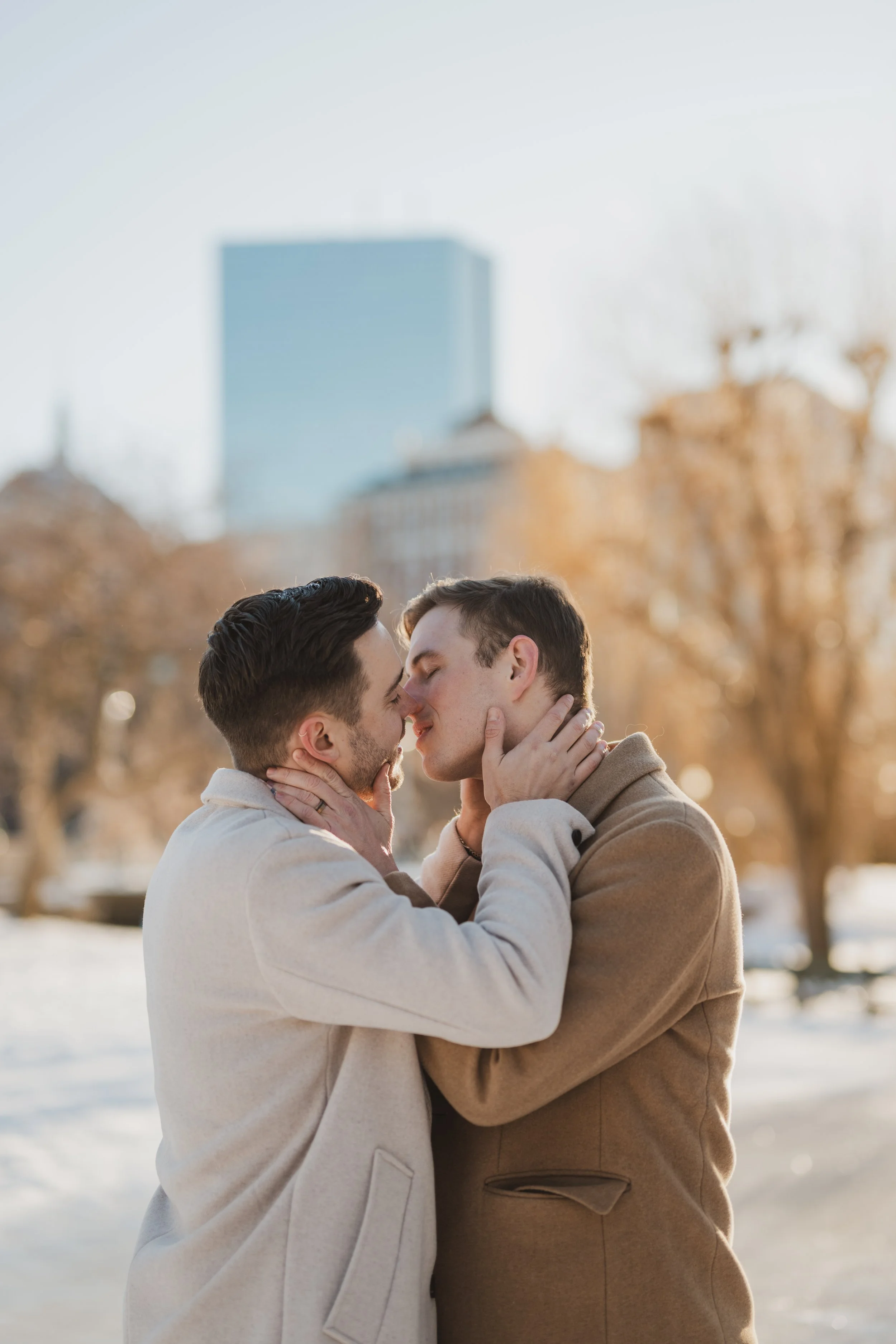 Two men stand in cream and brown pea coats on the ice covering the pond in Boston Common, holding each other's faces and about to kiss.