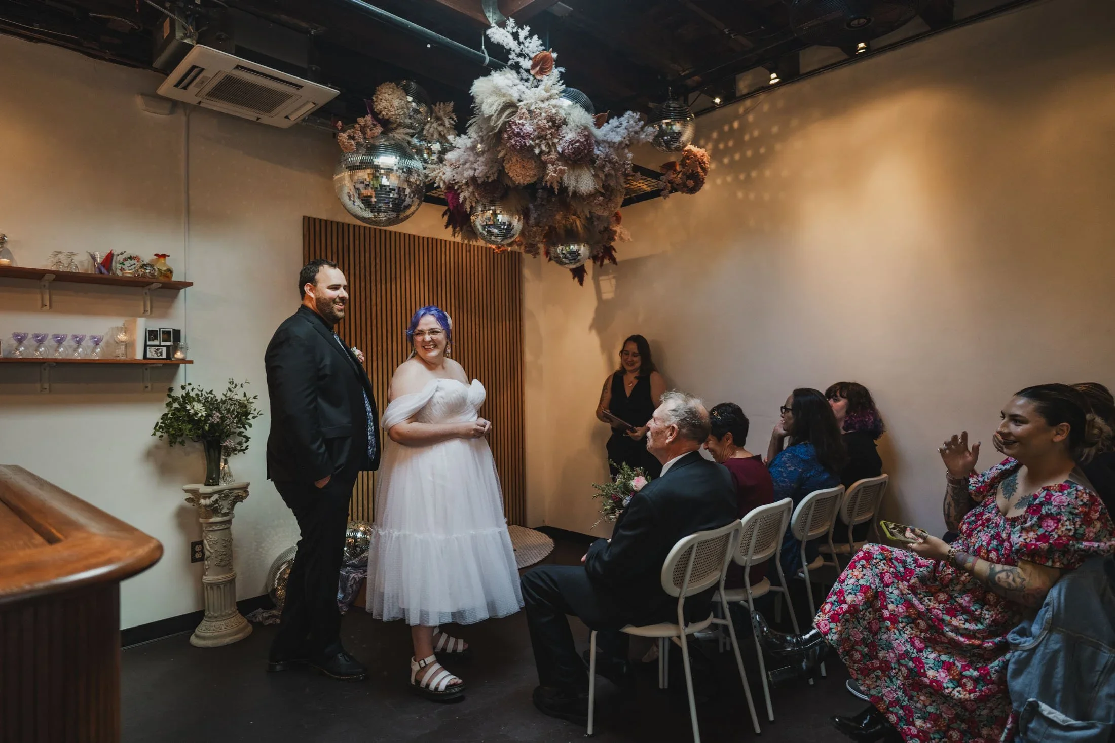 A couple stands, smiling looking around at their wedding guests in Dearly Studio shortly before the start of their ceremony