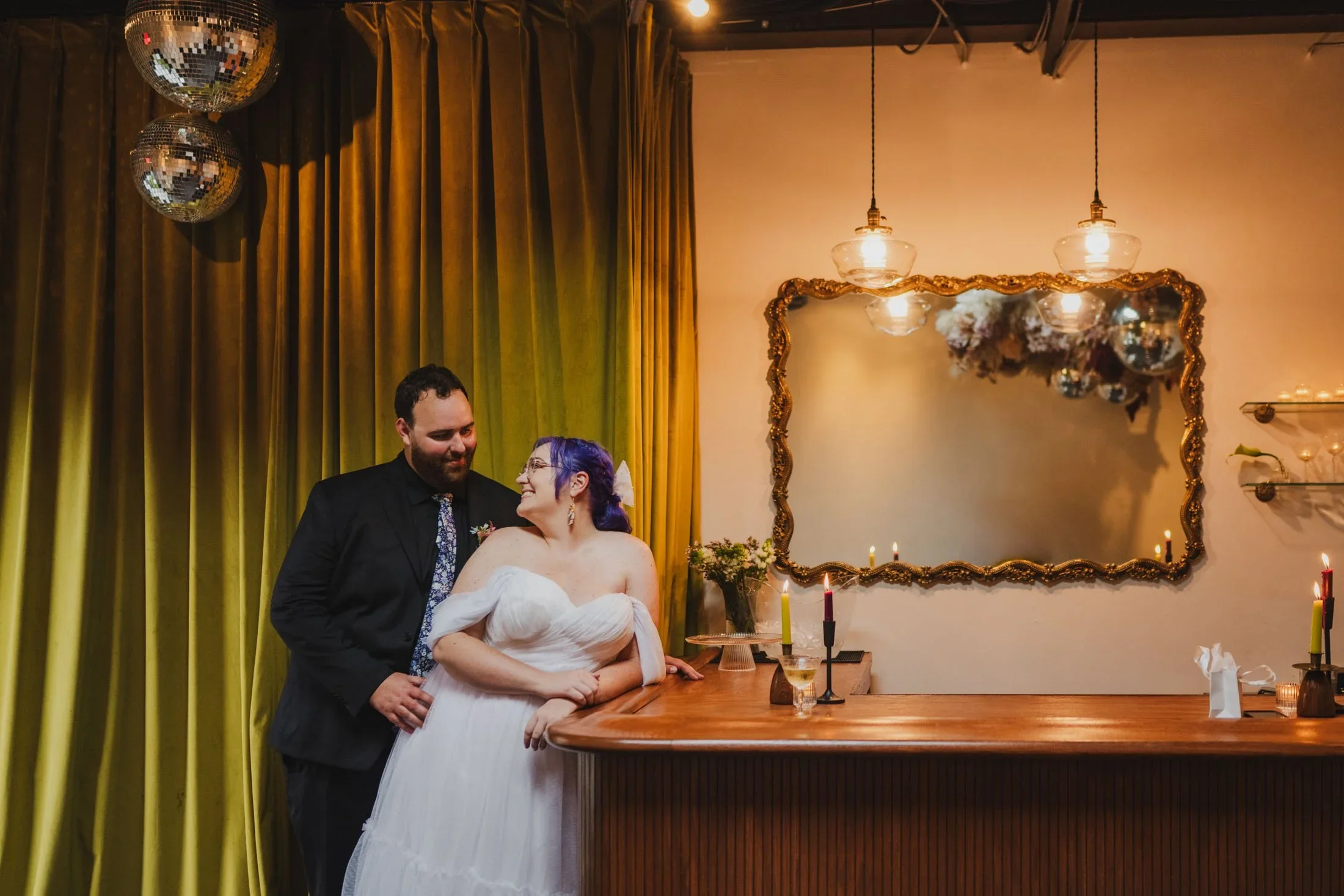 Quirky, stylish couple in wedding attire lean against the bar at Dearly Studio gazing at one another shortly before their ceeremony.