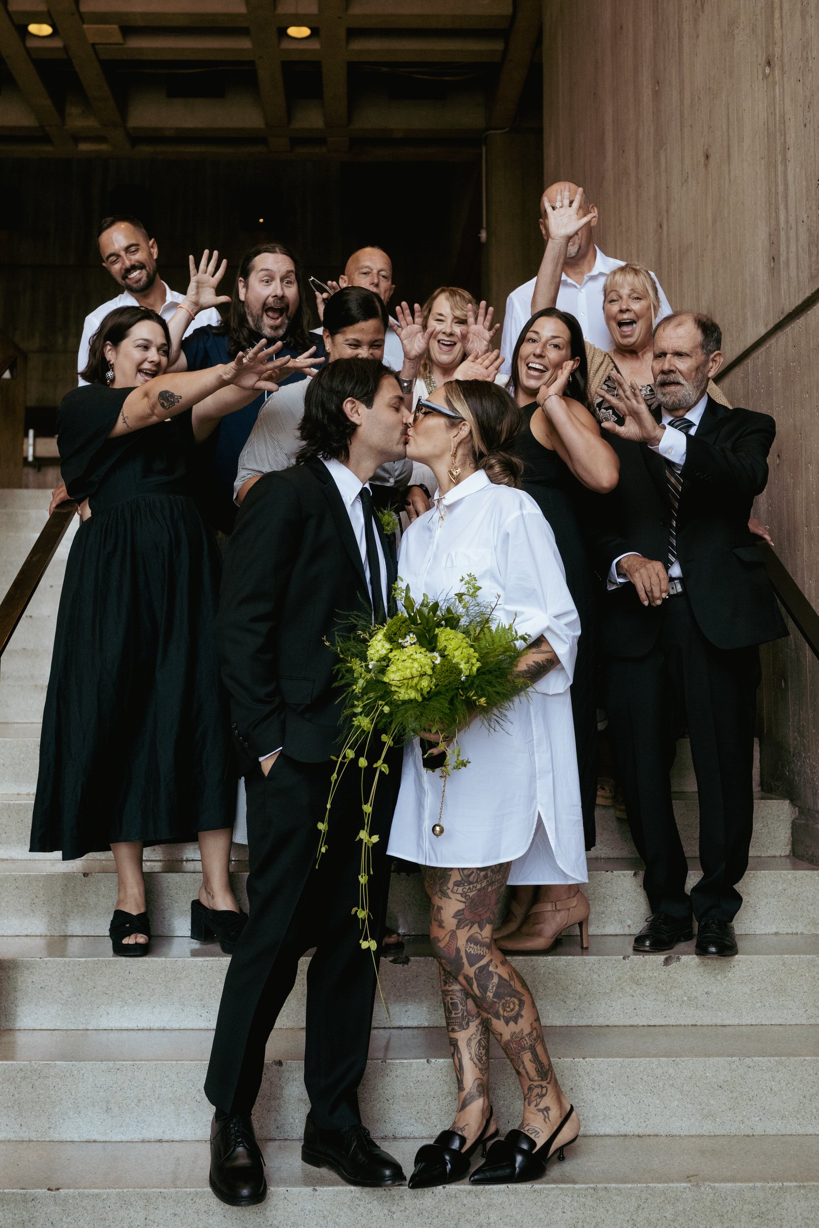 A newlywed couple kissing with their wedding guests celebrating behind them