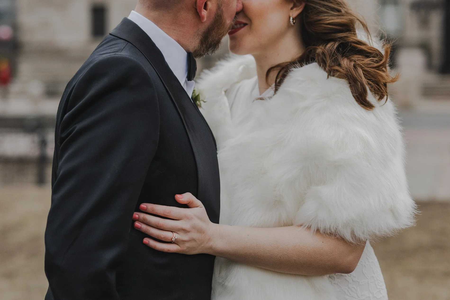 A couple stands in front of the Boston Public Library in winter about to kiss with the ring and fur shawl in focus.