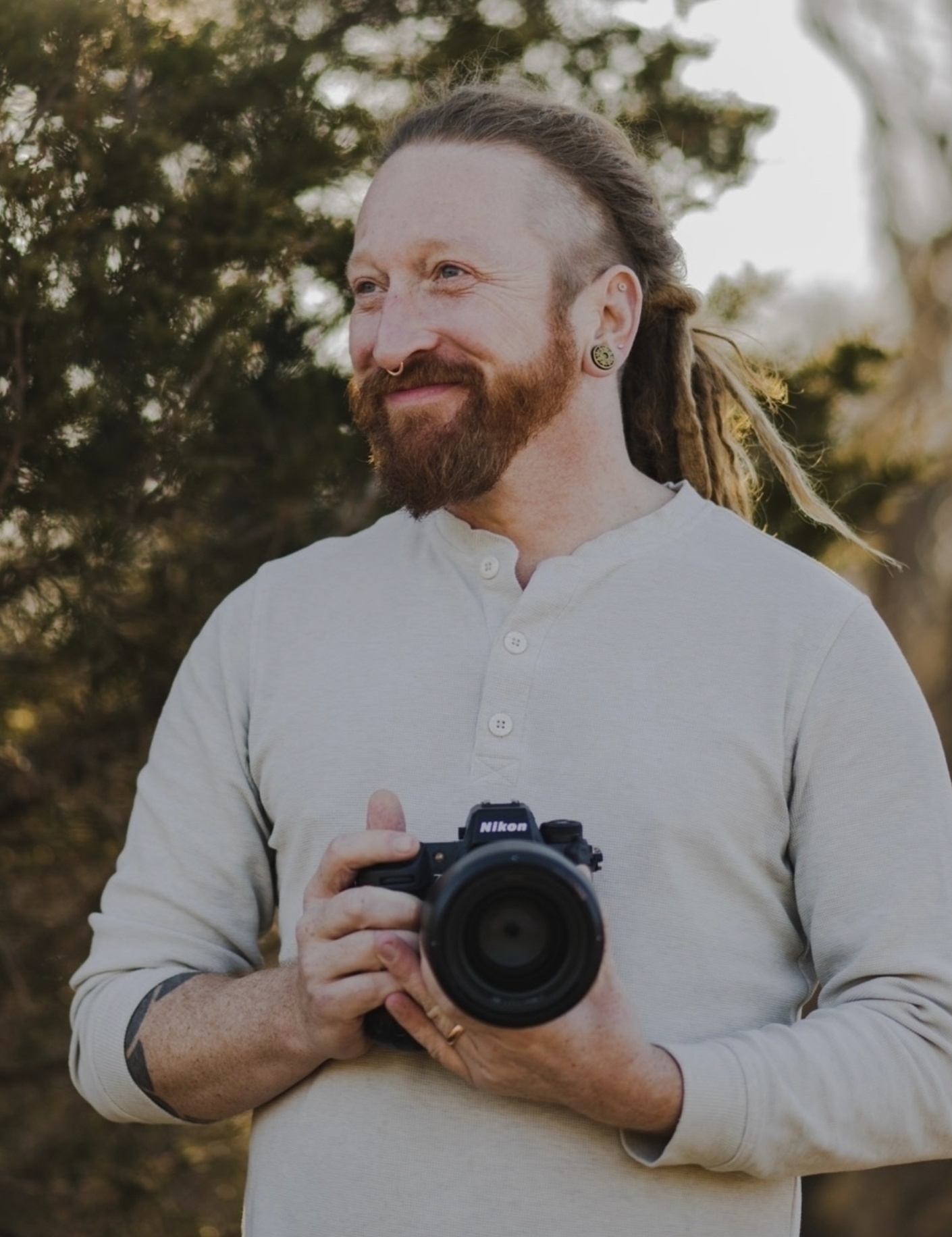 A smiling man with a red beard holding a Nikon looks off camera.