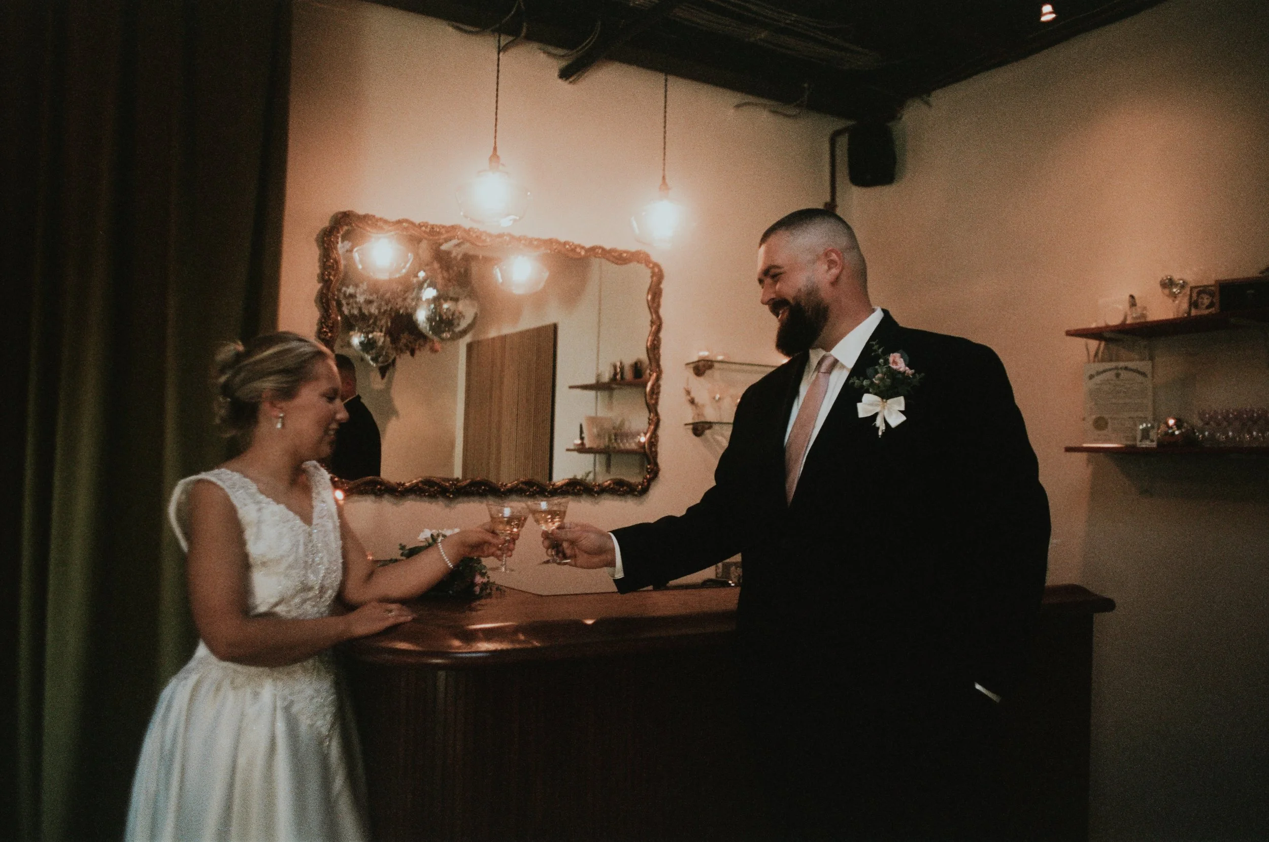 A 35mm film print of a couple in wedding attire leaning against a bar, clinking champagne coupes before saying their vows at Dearly Studio.