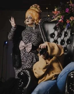 Amanda sitting in a chair with a camera with a drag queen standing next to her