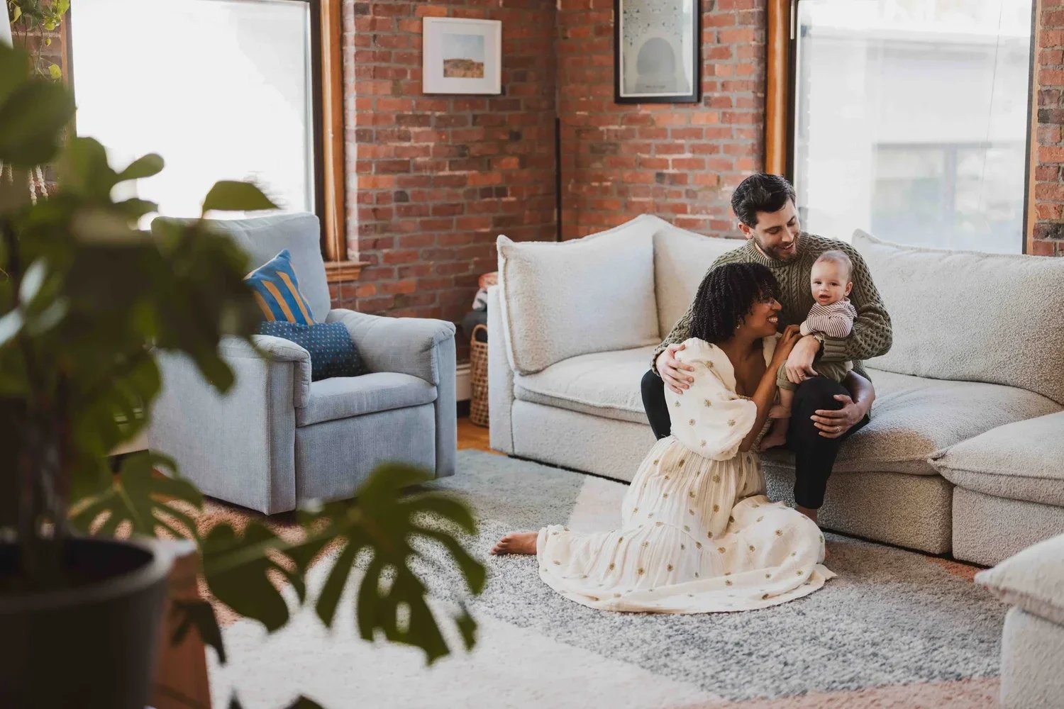 A family in their living room: the mother in a flowy cream dress sits on the floor between her husband’s legs as he holds their baby, and both parents smile at the child.