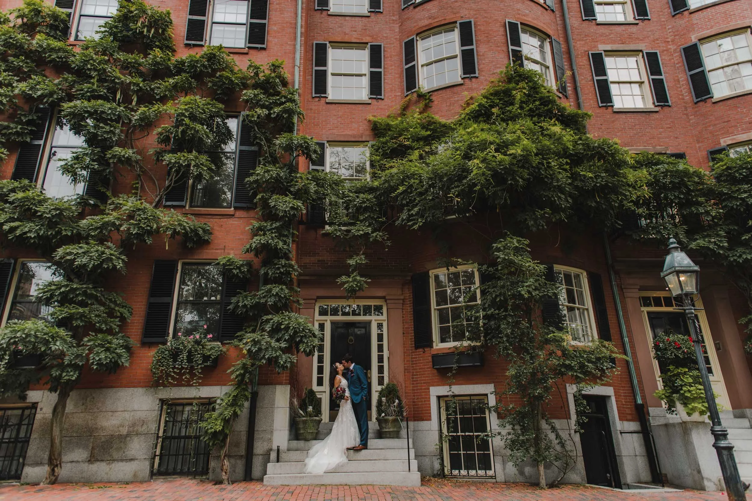 A newly married couple in wedding attire stands on a stoop in back bay surrounded by wisteria vines, kissing.