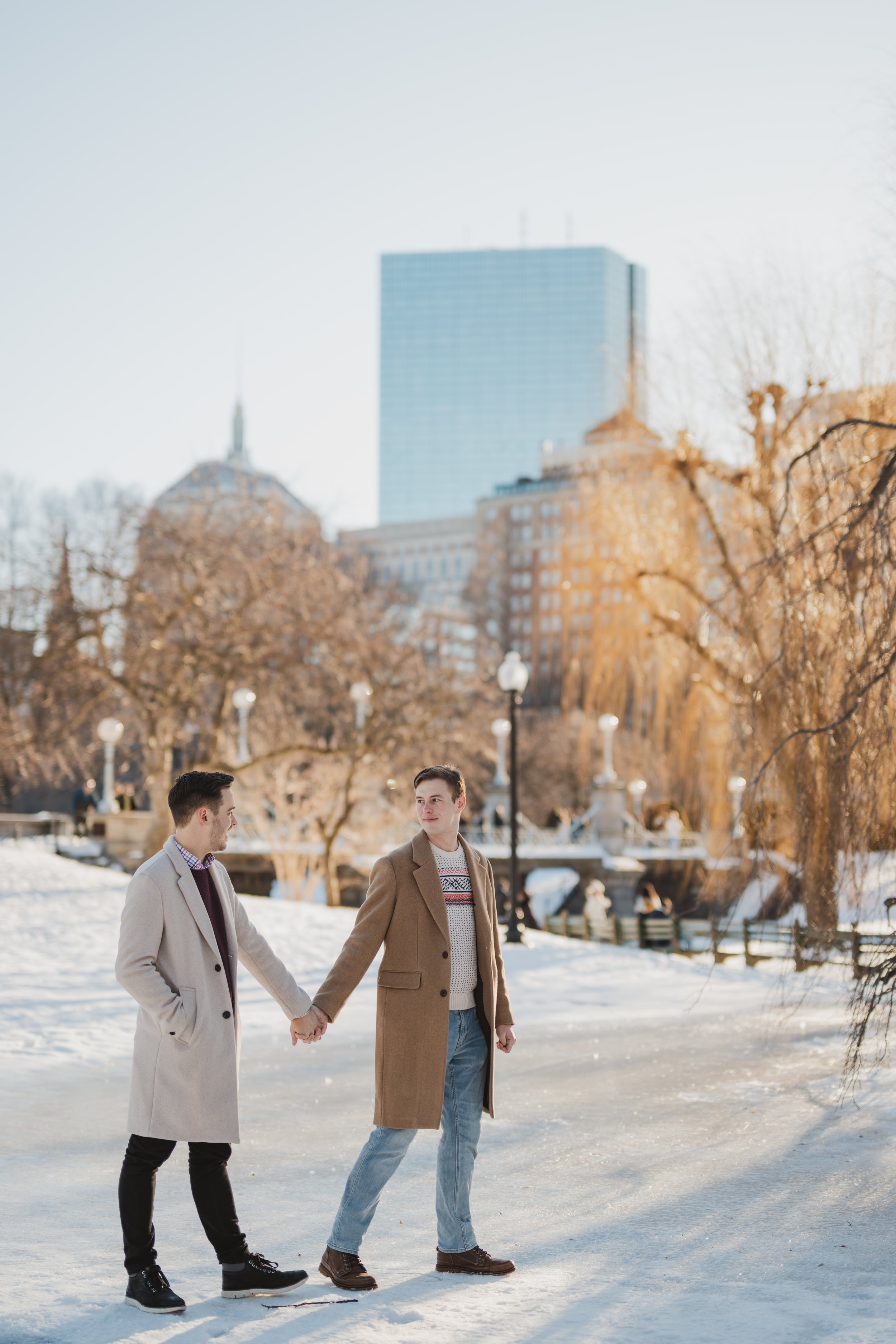 Two people stand hand-in-hand in a snowy Boston Public Garden with the historic bridge and city in the background as they stroll across the ice.