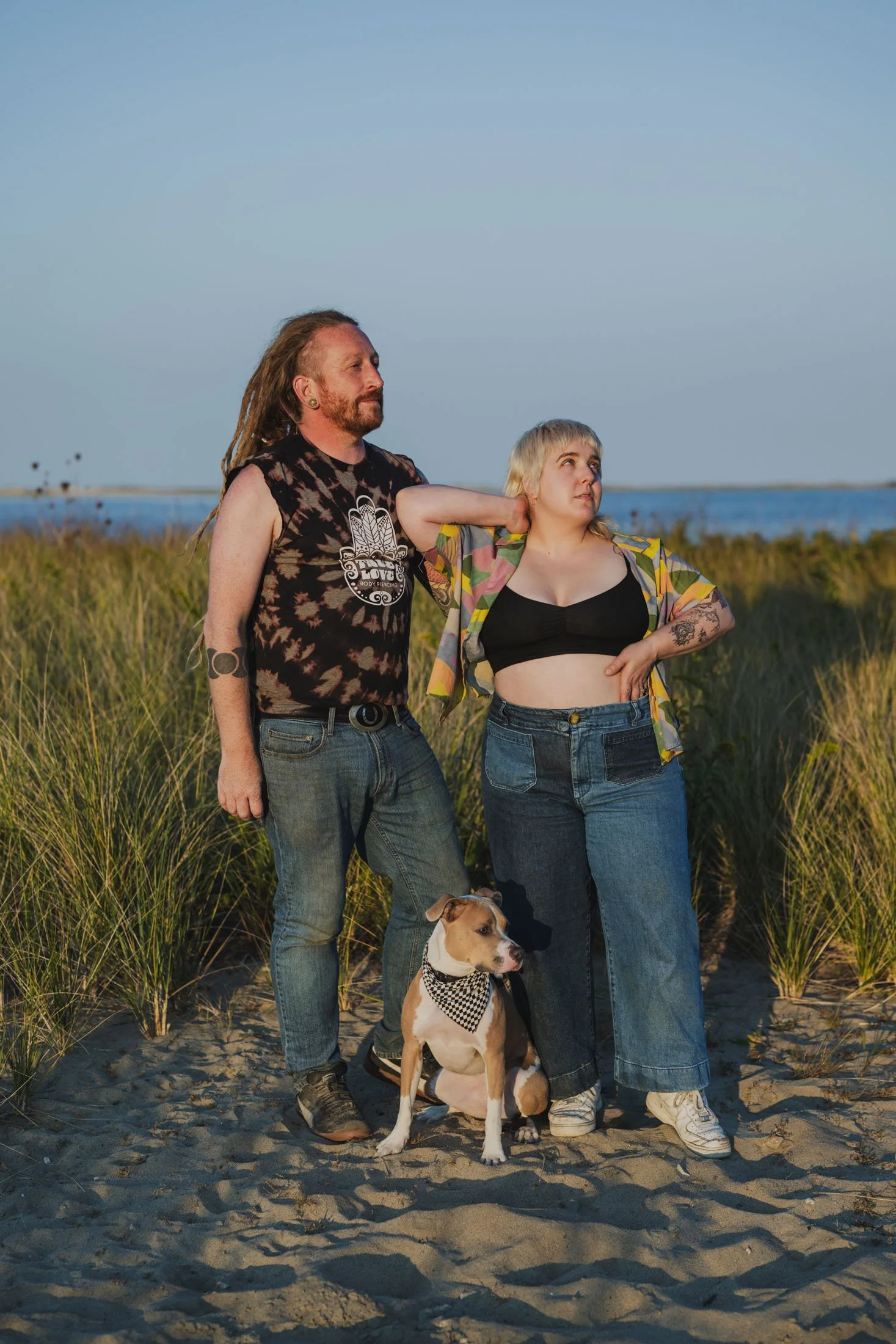 Owners of AMP are standing on a beach with their dog looking off into the sunset.