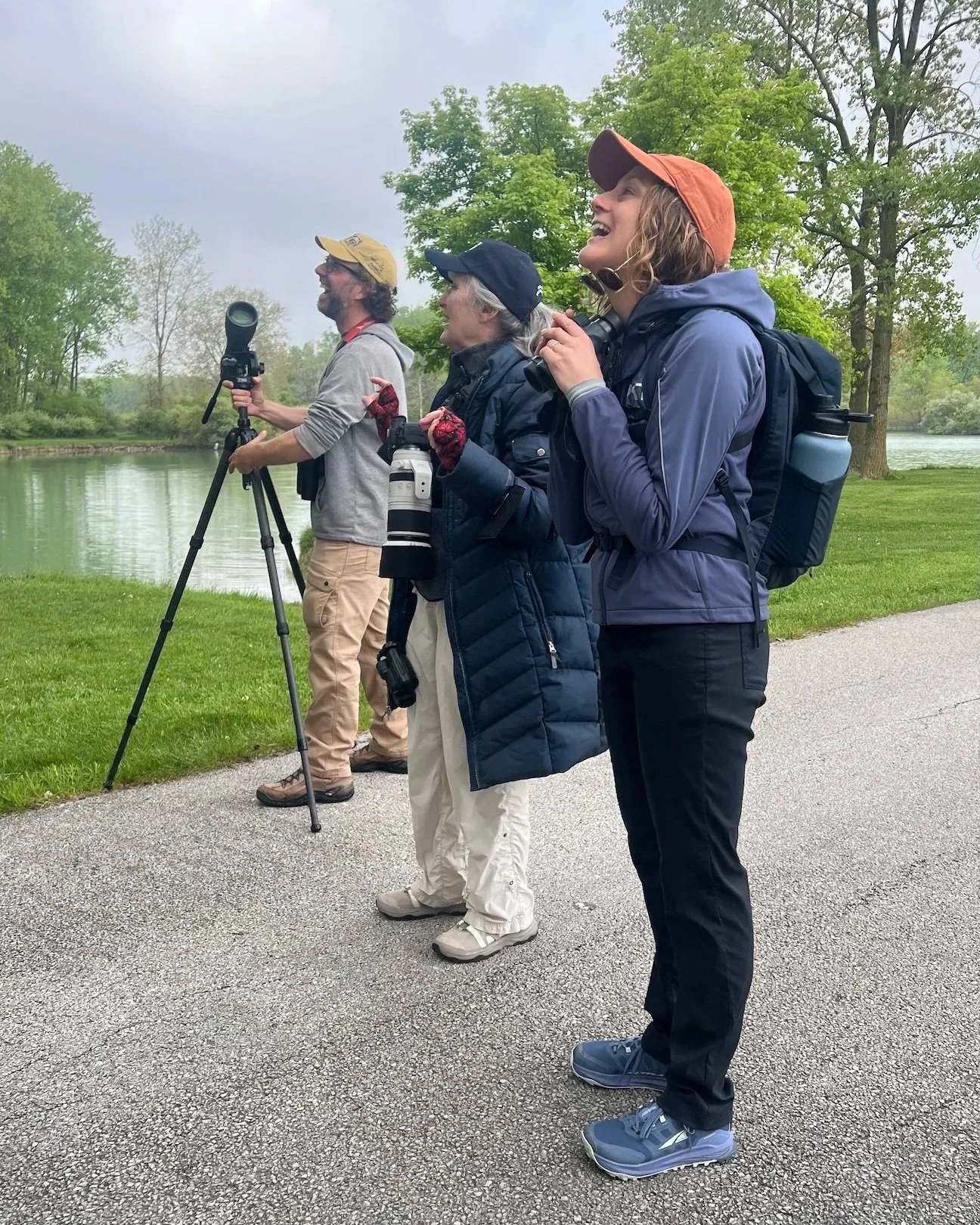 Three white birders laughing in unison. One Young bearded man, one older silver-haired woman and one young brown-haired woman. All hold optical euipment and wear baseball caps.