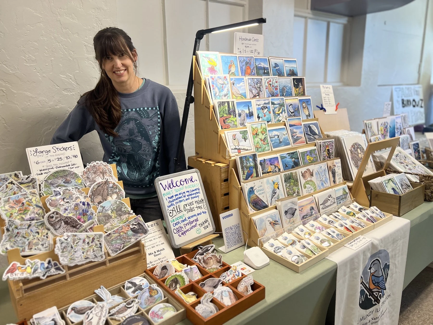 Woman standing behind a table filled with greeting cards and stickers with signage indicating prices. Woman is smiling at the photographer.