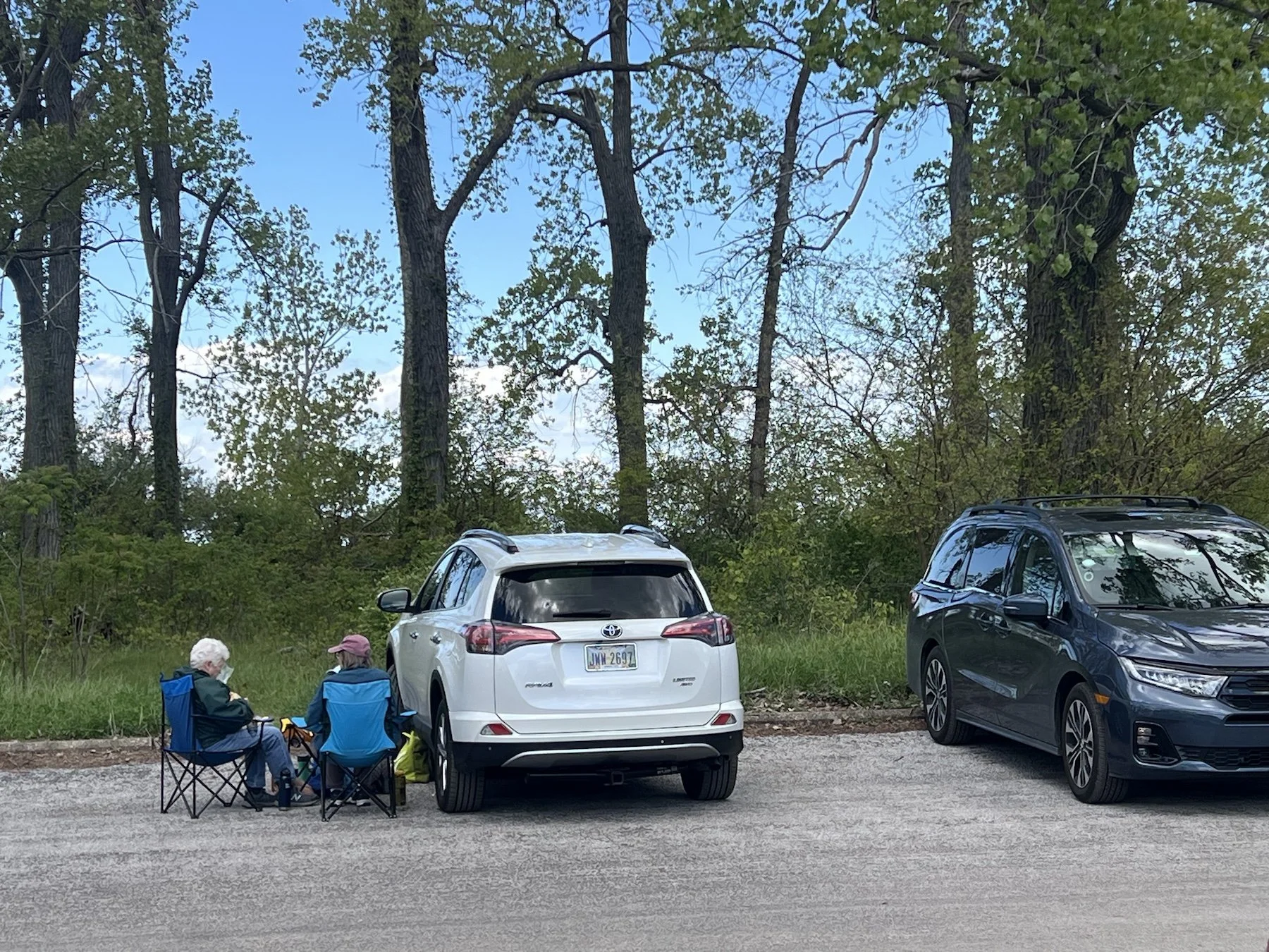 Two women sitting in camp chairs beside their parked car, ina tree lined parking lot.