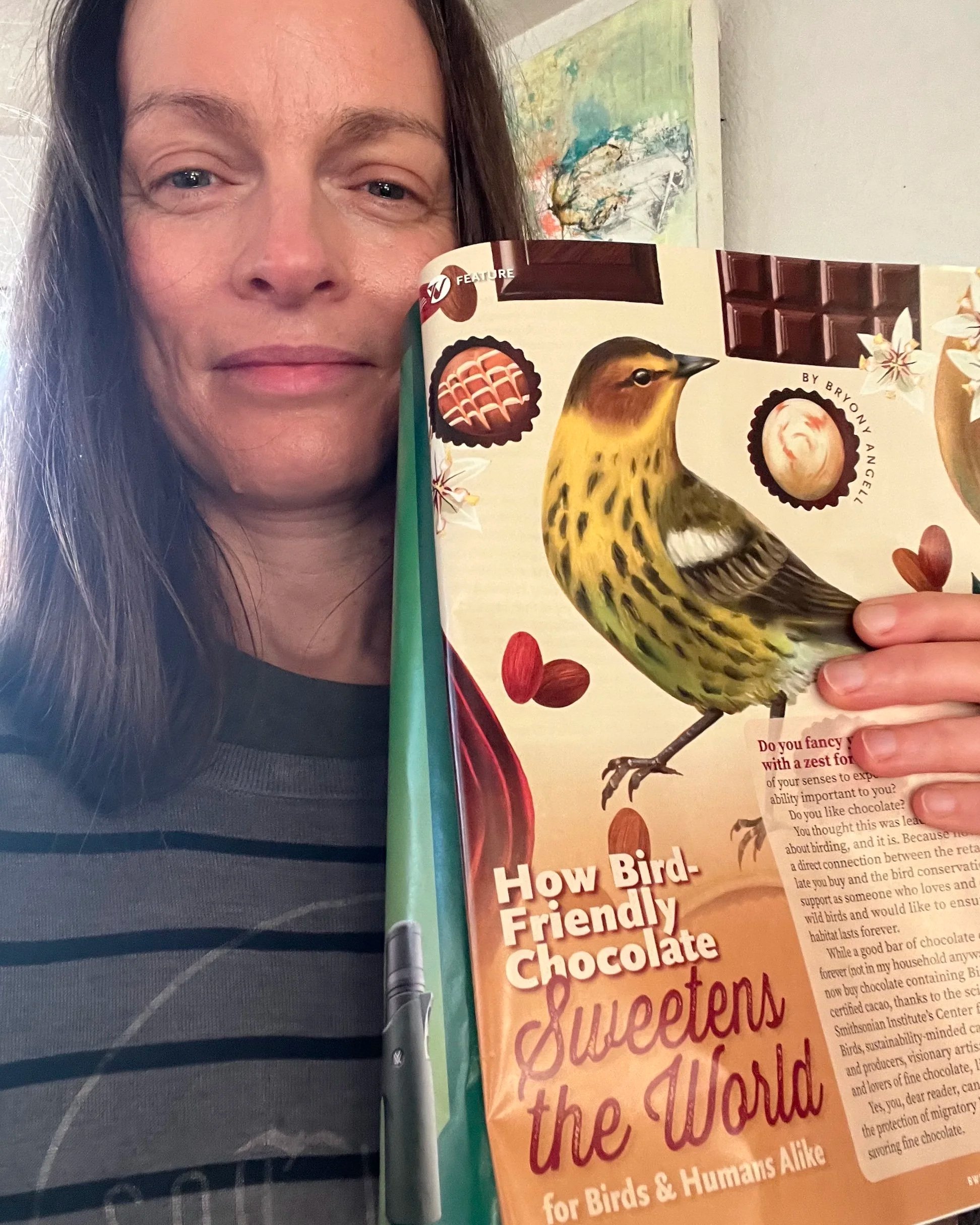 Woman holding a magazine with the pages showing an image of a bird and chocolates. The page is open to show the title of the article, "How Bird Friendly Chocolate Sweetens the World for Birds and Humans Alike."