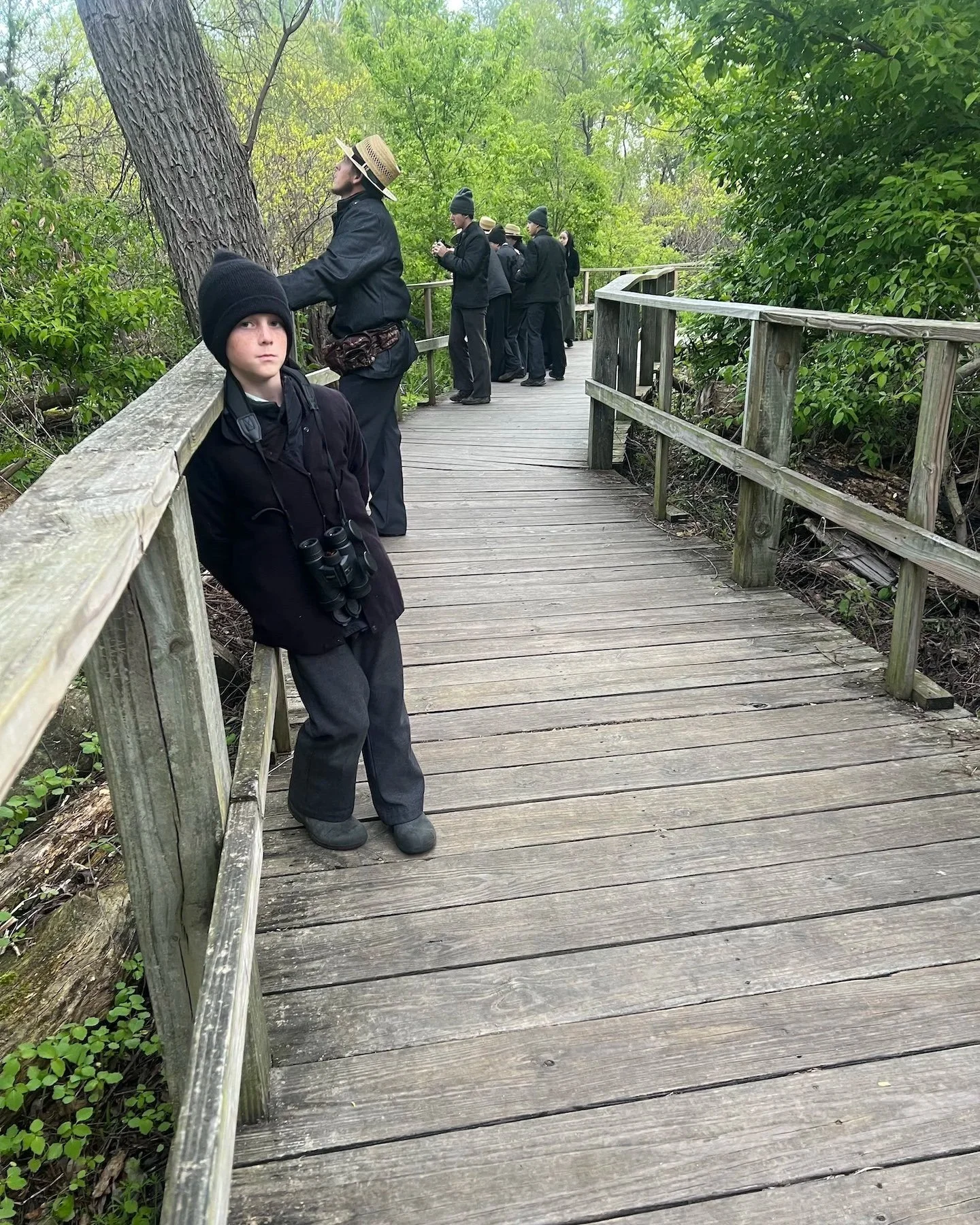 Young boy looking directly at the camera while holding binoculars. Boy stands on a boardwalk in a marsh, with family members in the back ground.