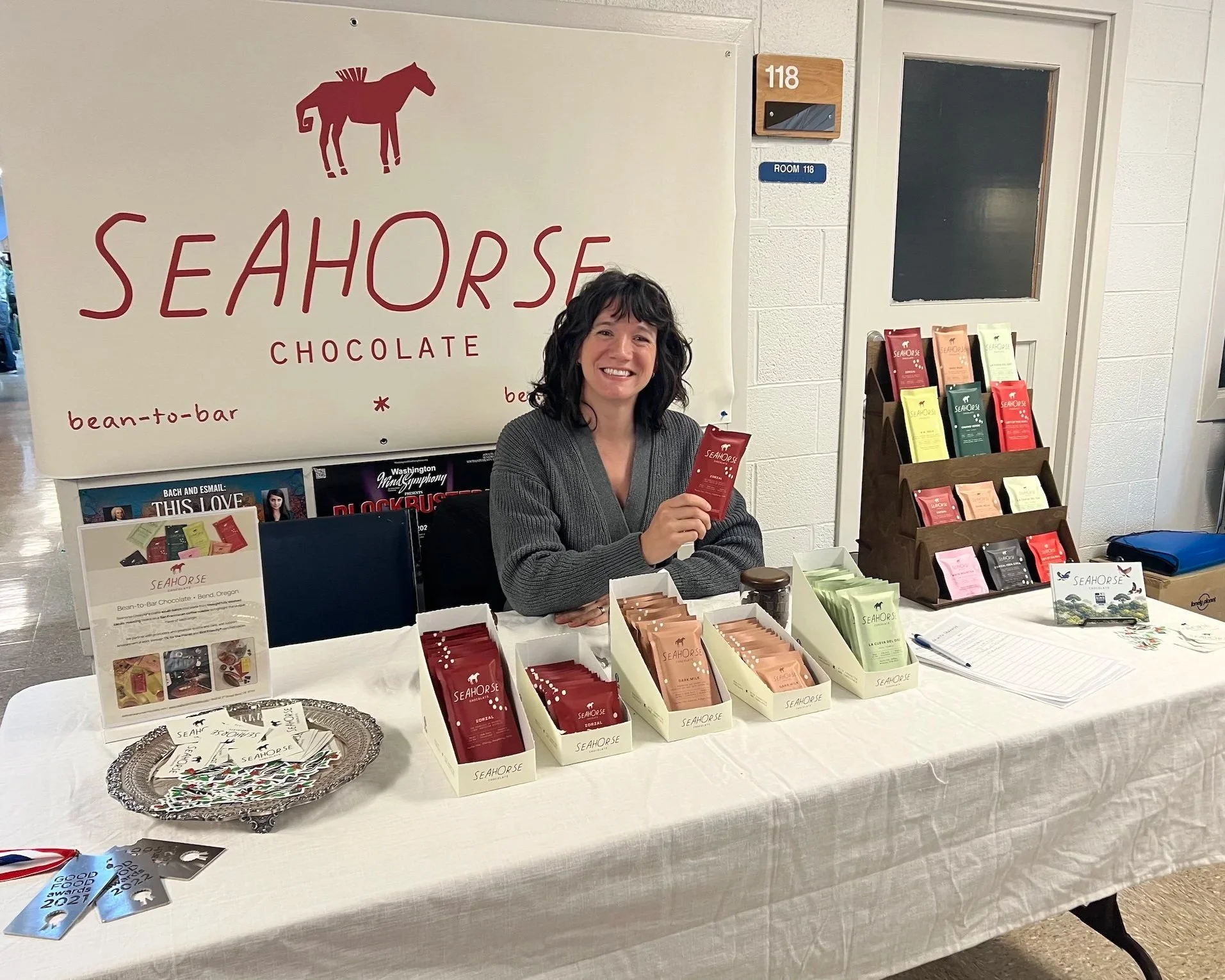 Smiling woman sitting behind a table displaying Seahorse Chocolate brand items. Woman is holding a packaged chocolate bar up for the camera.