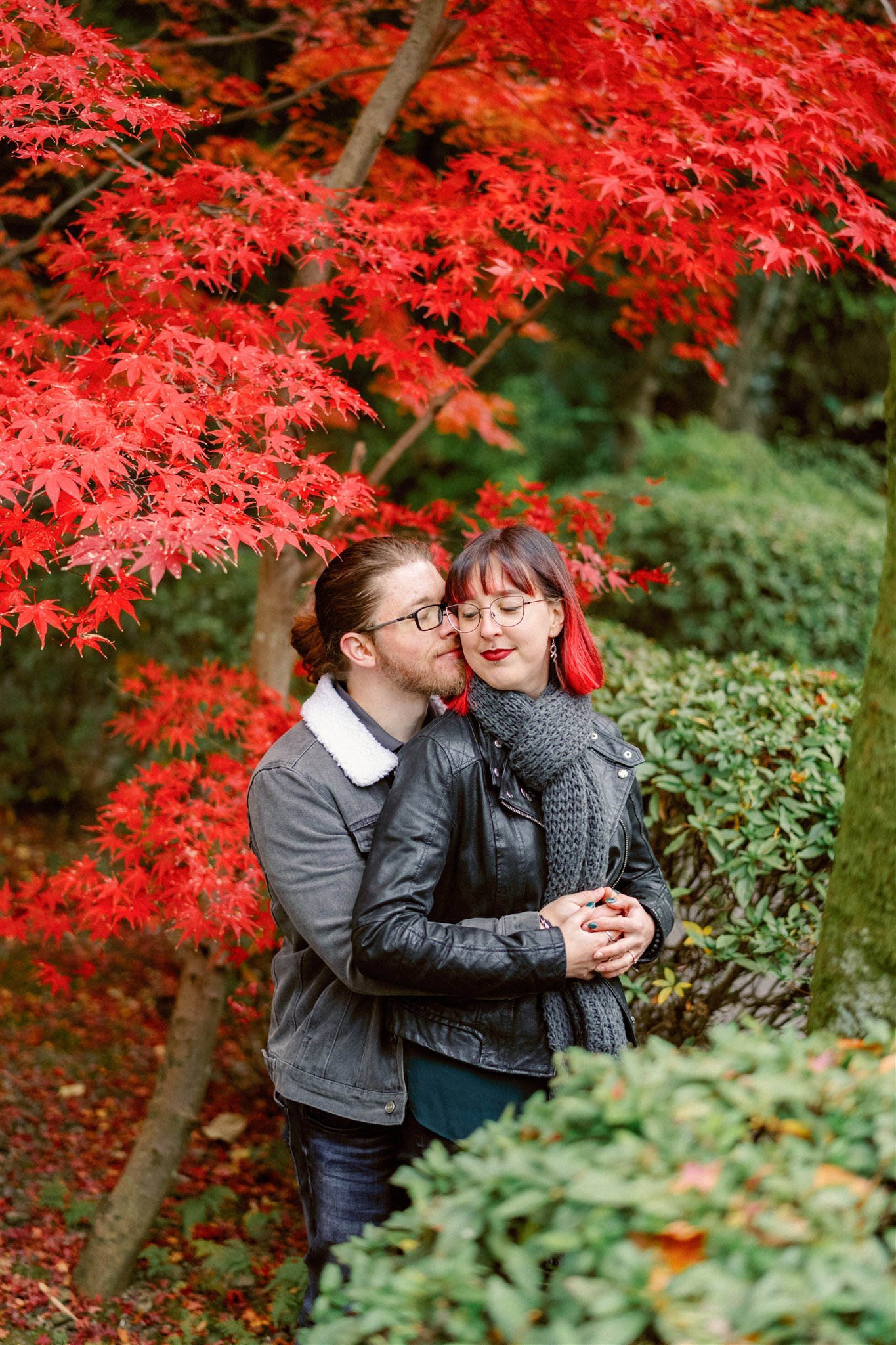 Couple hug during their engagement photoshoot in front of vibrant red autumn leaves.
