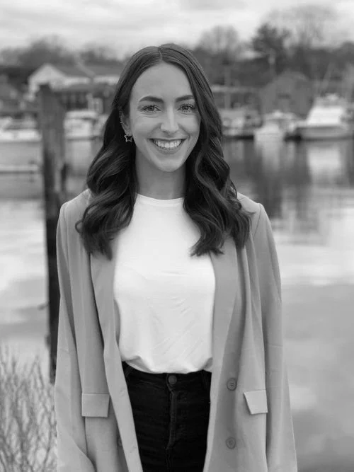 A woman smiling outdoors near a body of water with boats in the background, wearing a white shirt and a light-colored blazer.