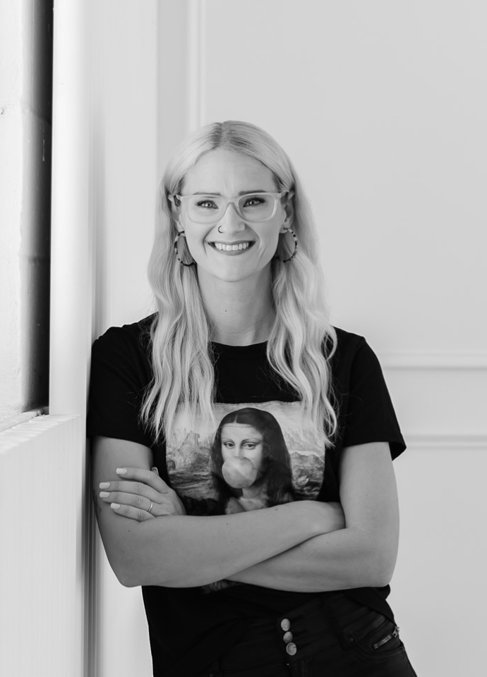 A young woman with long blonde wavy hair, glasses, and hoop earrings, smiling and crossing her arms, wearing a T-shirt featuring the Mona Lisa and a bubblegum blowing girl, standing next to a wall.