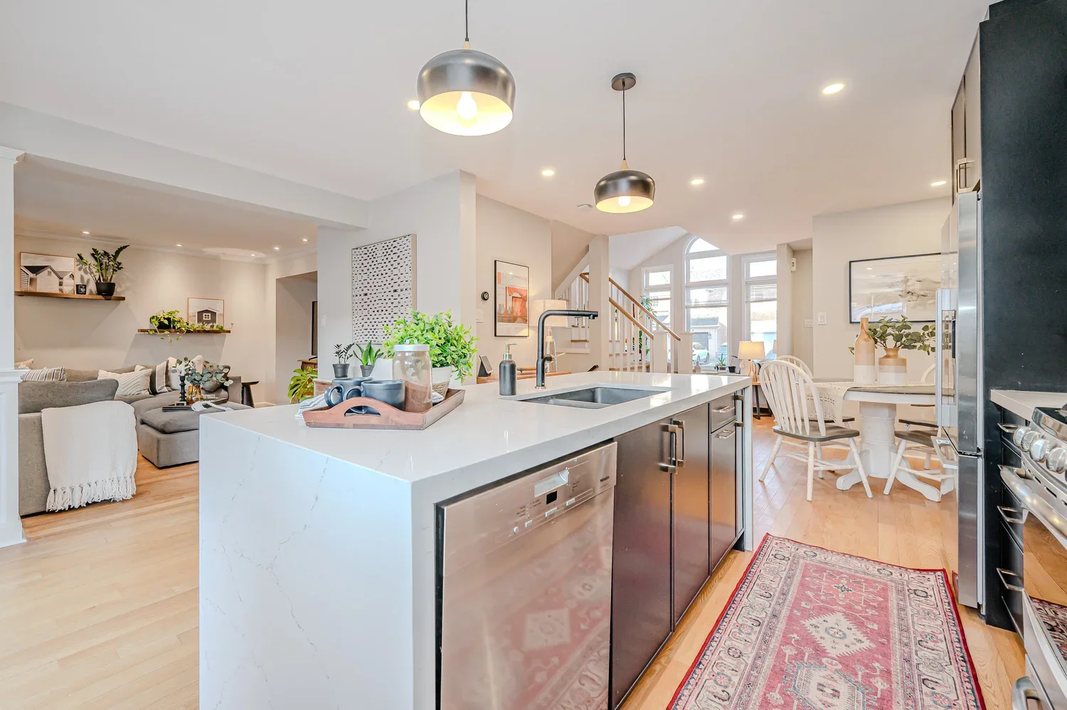 Open-concept kitchen and living room with white walls, hardwood floors, and modern decor. The kitchen has a white countertop, black cabinets, and stainless steel appliances. There are pendant lights above the island, which features a sink and a toaster. The living room has a gray sectional sofa, bookshelves, and wall art, with natural light coming through large windows at the back.