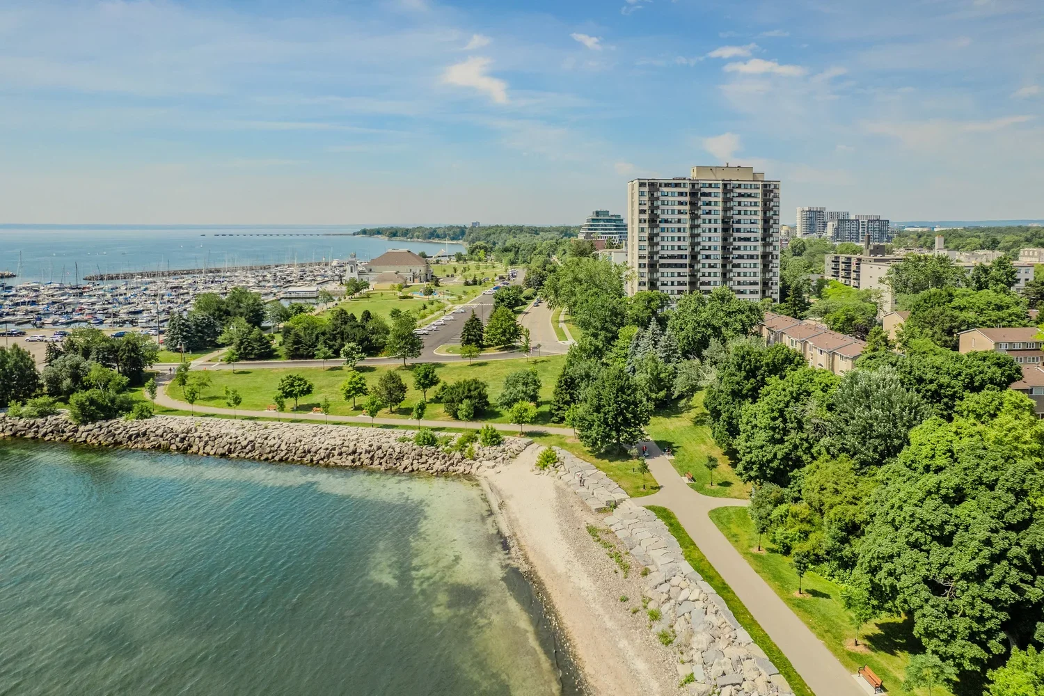 A coastal cityscape featuring a marina filled with boats, a park with walking paths and green trees, and high-rise buildings in the background alongside residential houses. The shoreline is lined with rocks and a sandy beach, under a partly cloudy sky.