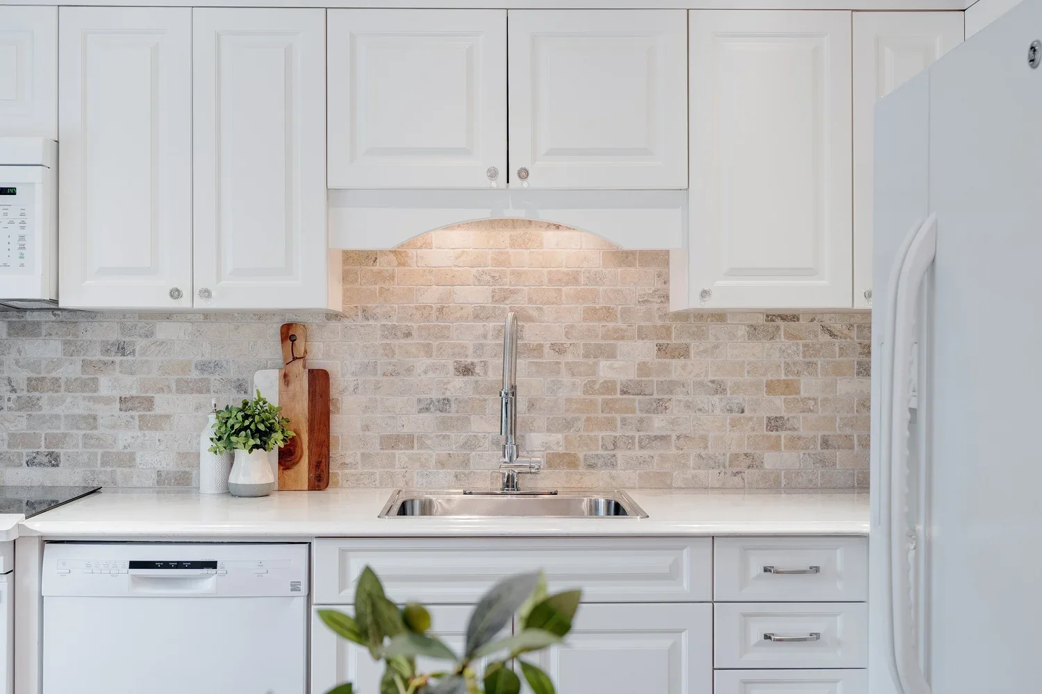 White kitchen with upper cabinets, a brick backsplash, a stainless steel sink, a dishwasher, and a refrigerator. Decor includes a potted plant, a cutting board, and a paper towel roll.