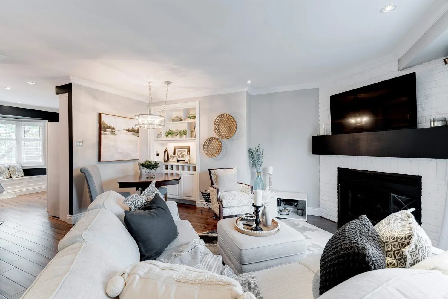 Living room with white walls, wooden flooring, a fireplace with a mounted TV, a beige sofa with black and beige pillows, a white ottoman with candles, a armchair with a pillow, a collage of baskets and a vase with greenery, a dining area with a round table, and a window seat.