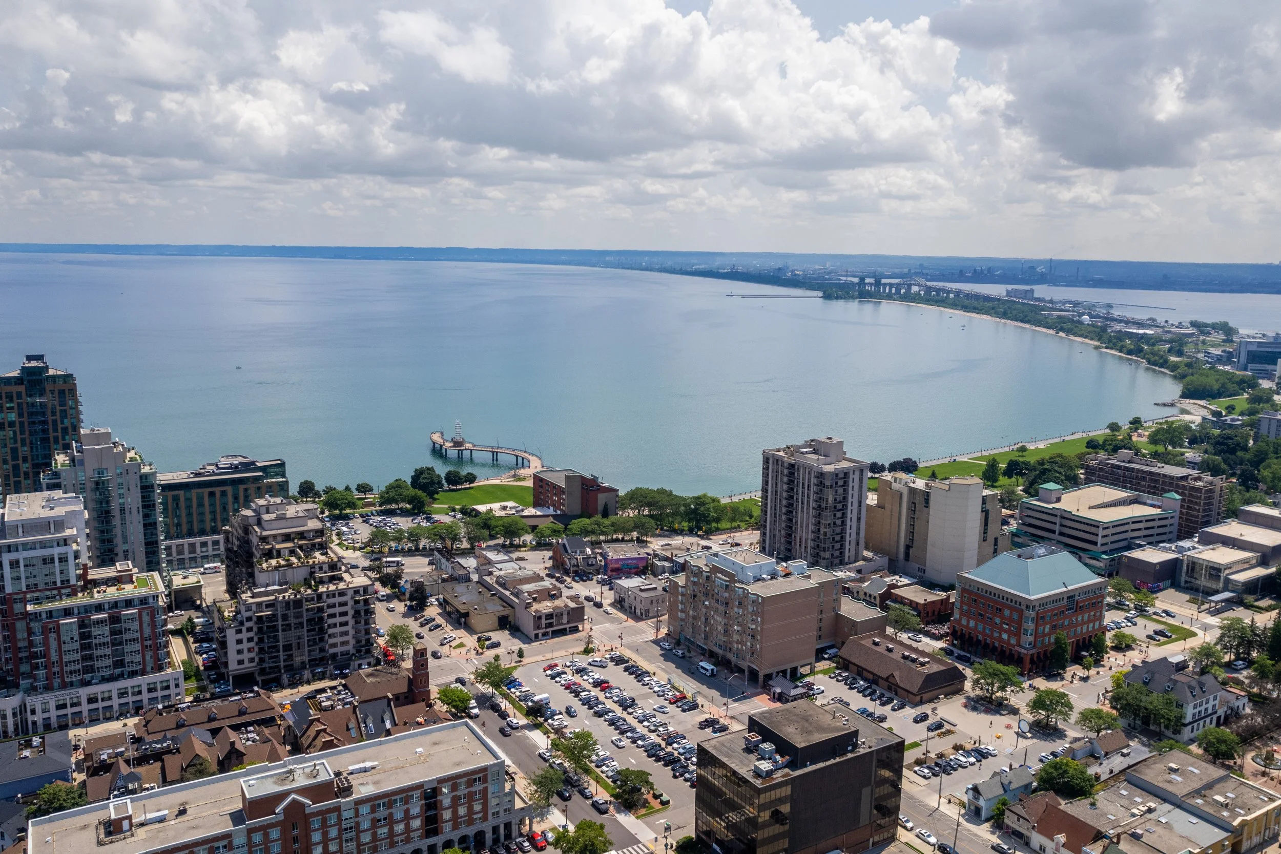 Aerial view of a lakeside cityscape with tall buildings, a park along the lake, and a pier extending into the water. Cloudy sky overhead.