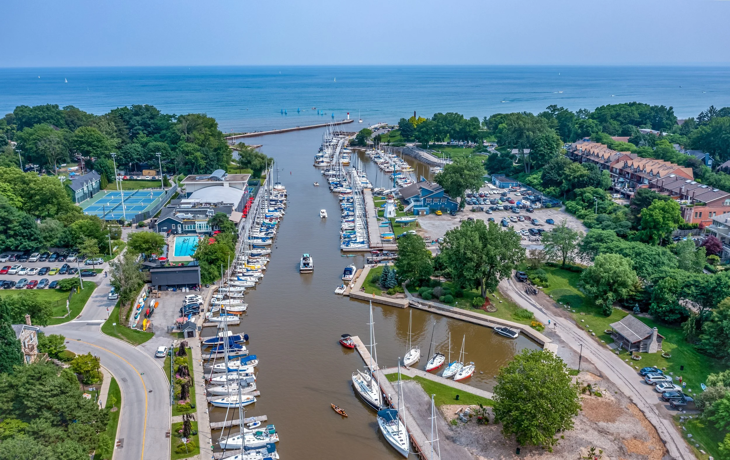 Aerial view of a marina with boats docked along a canal, surrounded by green trees, tennis courts, parking lots, residential buildings, and a body of water with sailboats.