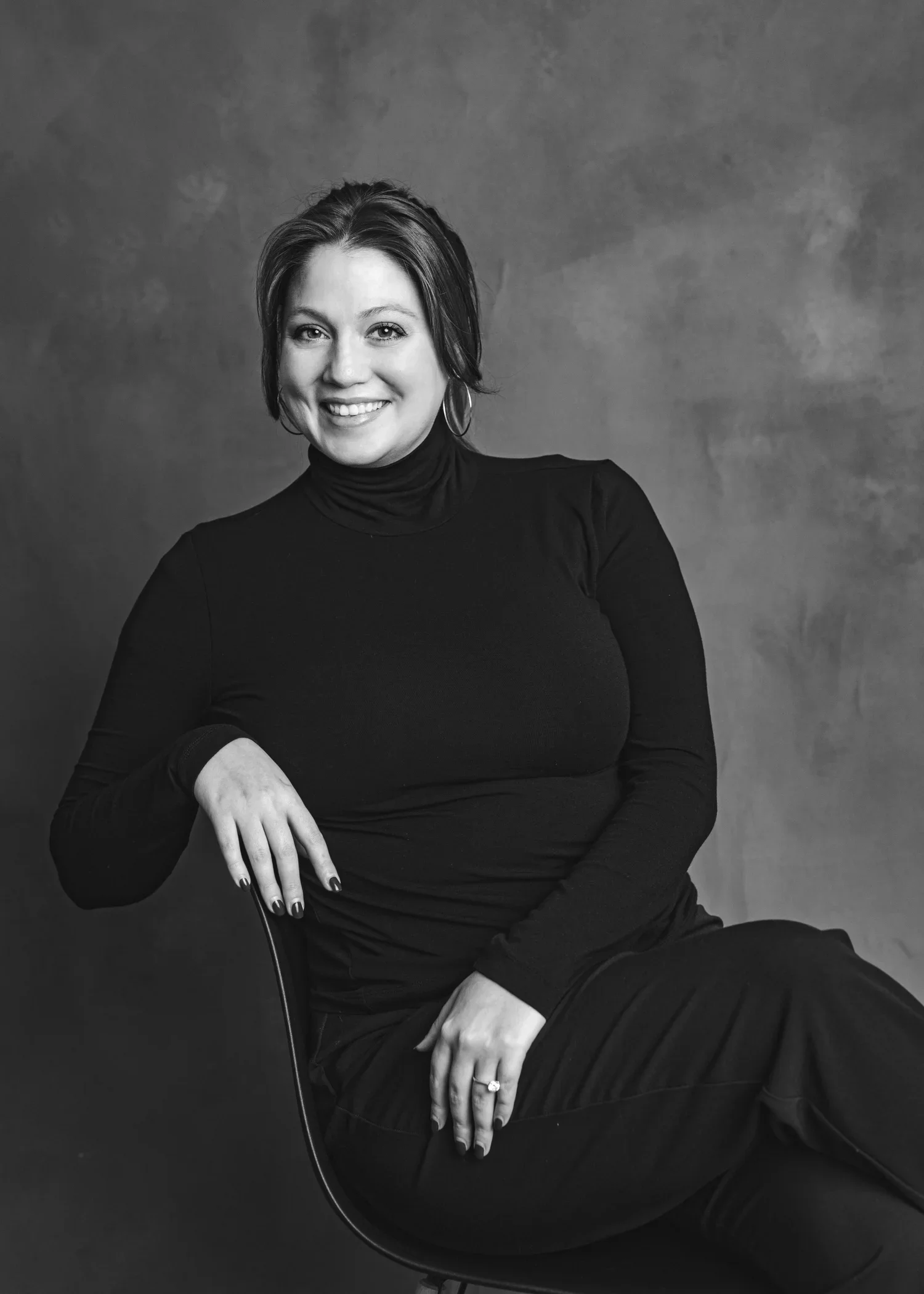 Black and white portrait of a woman smiling, wearing a turtleneck sweater and hoop earrings, sitting on a chair, against a plain background.