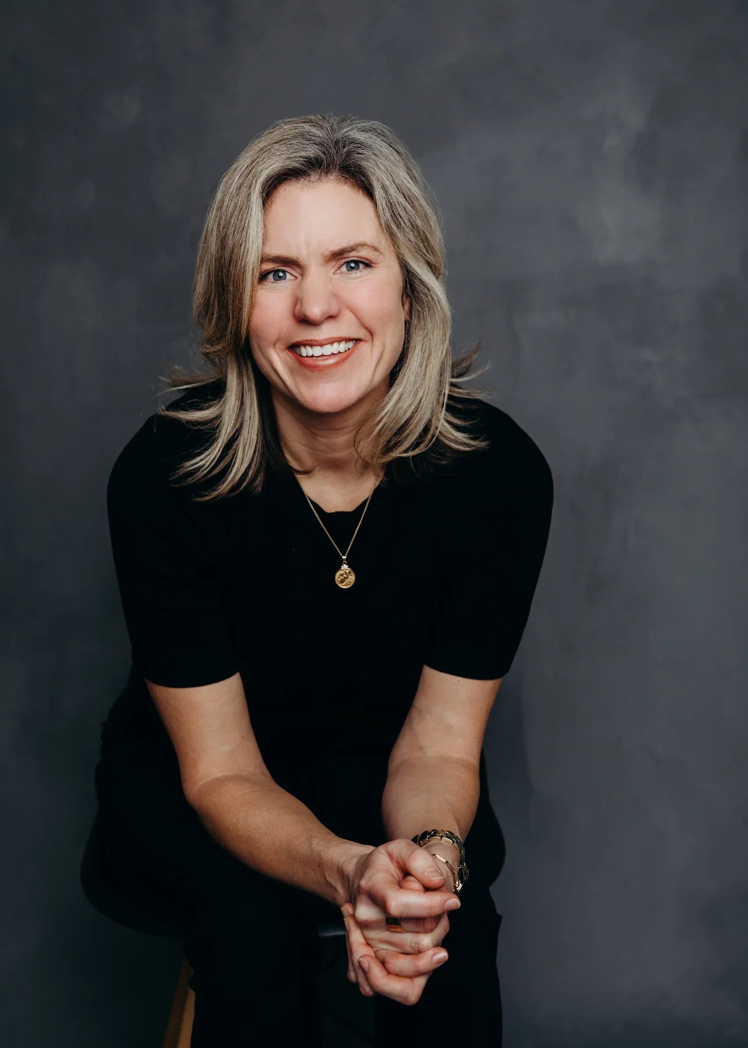 A woman with shoulder-length gray hair smiling, wearing a black top, gold necklace, and bracelets, sitting against a dark gray background.