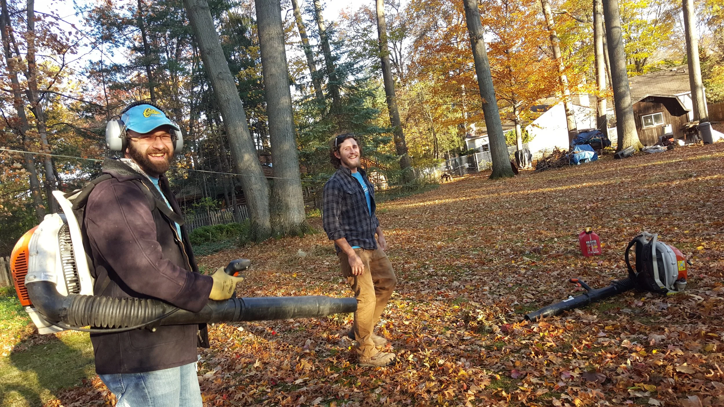 Two men smiling outdoors in a wooded area during fall, one holding a leaf blower, and the other standing among fallen leaves with a leaf blower on the ground nearby.